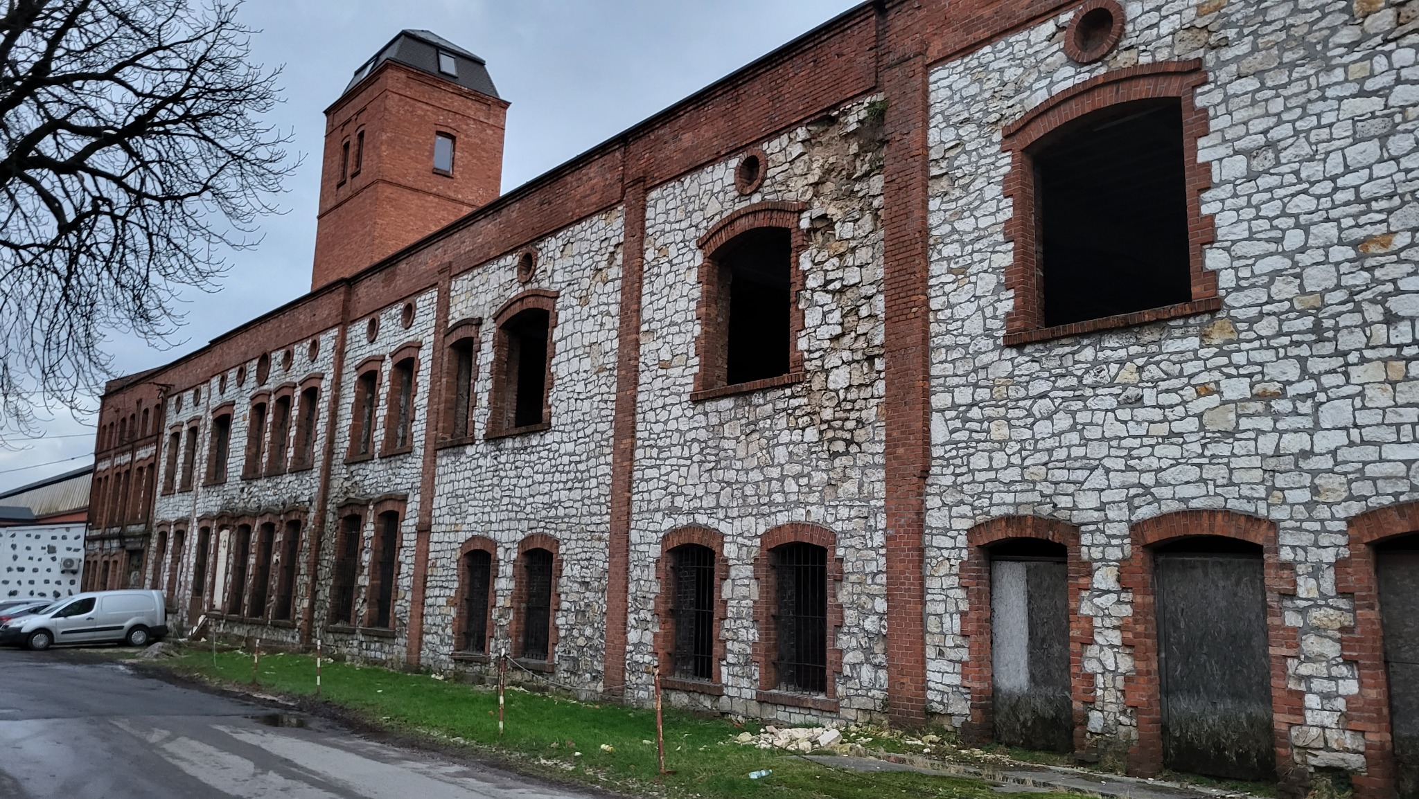 Długi, kamienno-ceglany budynek z wieżą w trakcie przeglądu okresowego, z widocznymi śladami zaniedbania i brakiem okien w niektórych otworach okiennych.