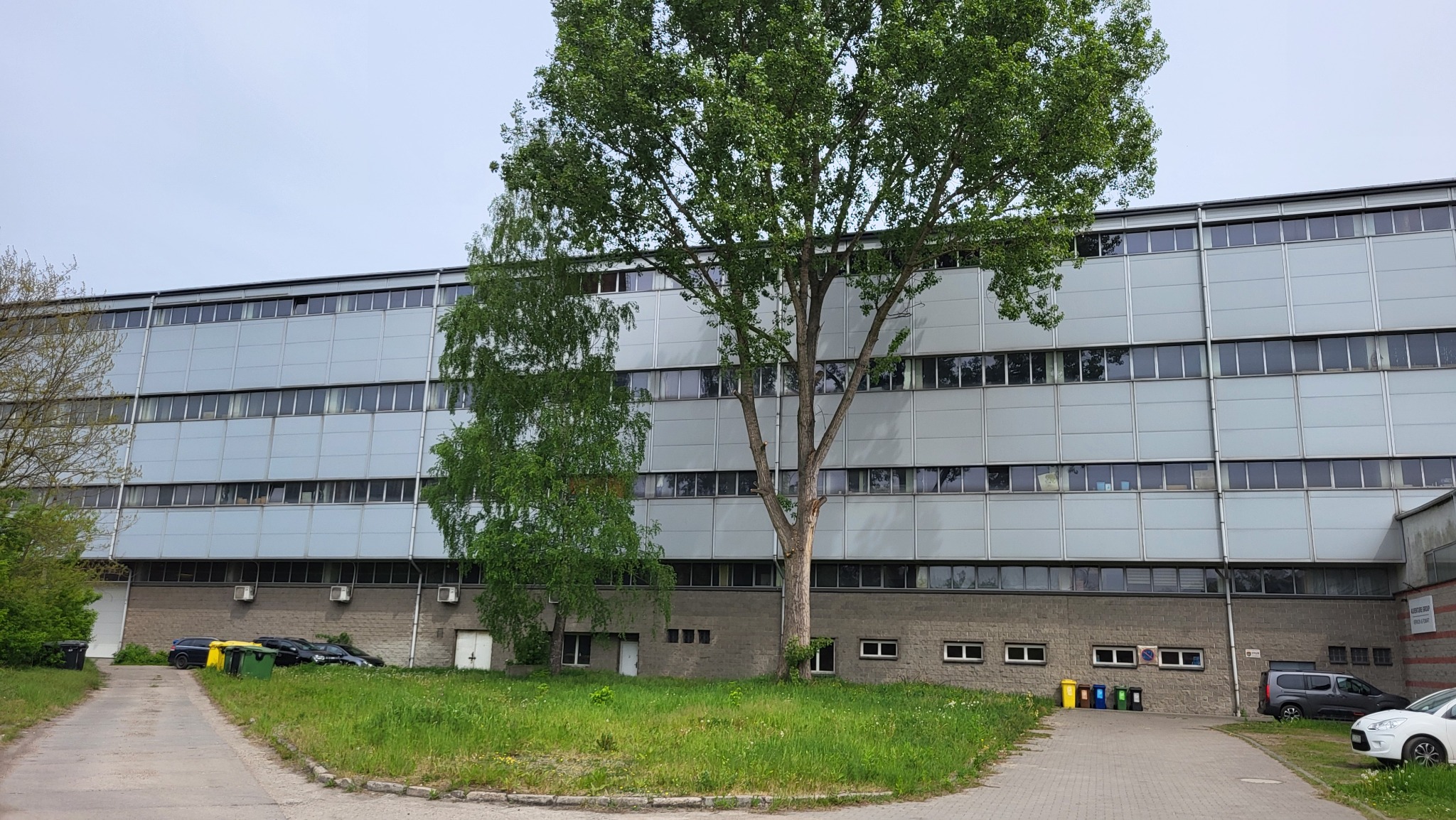 Wielopiętrowy budynek przemysłowy z licznymi oknami, widoczny z poziomu gruntu, z drzewami na pierwszym planie i kilkoma samochodami w tle.