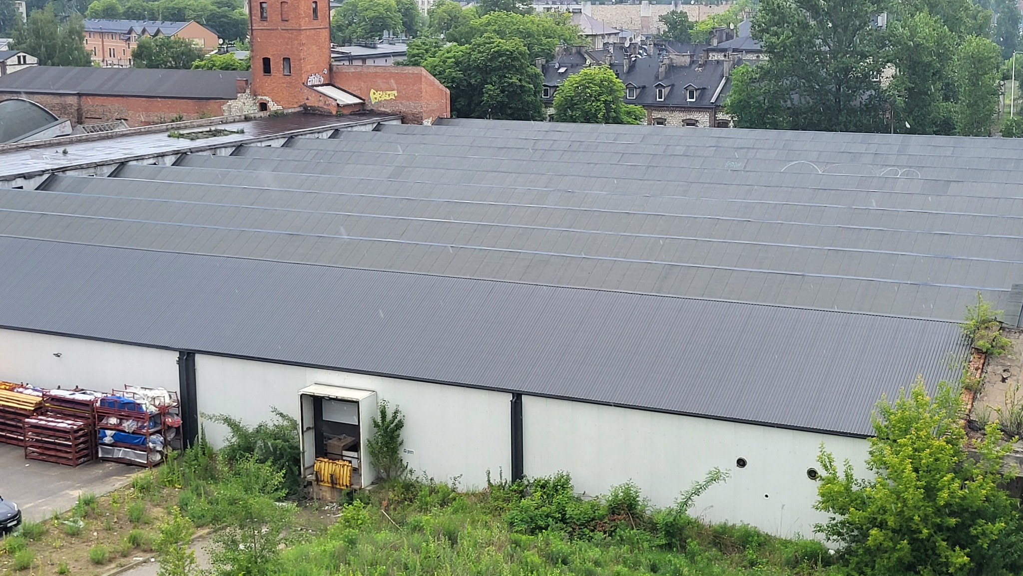 Widok z góry na duży budynek przemysłowy z szarym, falistym dachem, białą elewacją i otwartą bramą załadunkową, otoczony zielenią i z widoczną starą, ceglaną wieżą w tle.