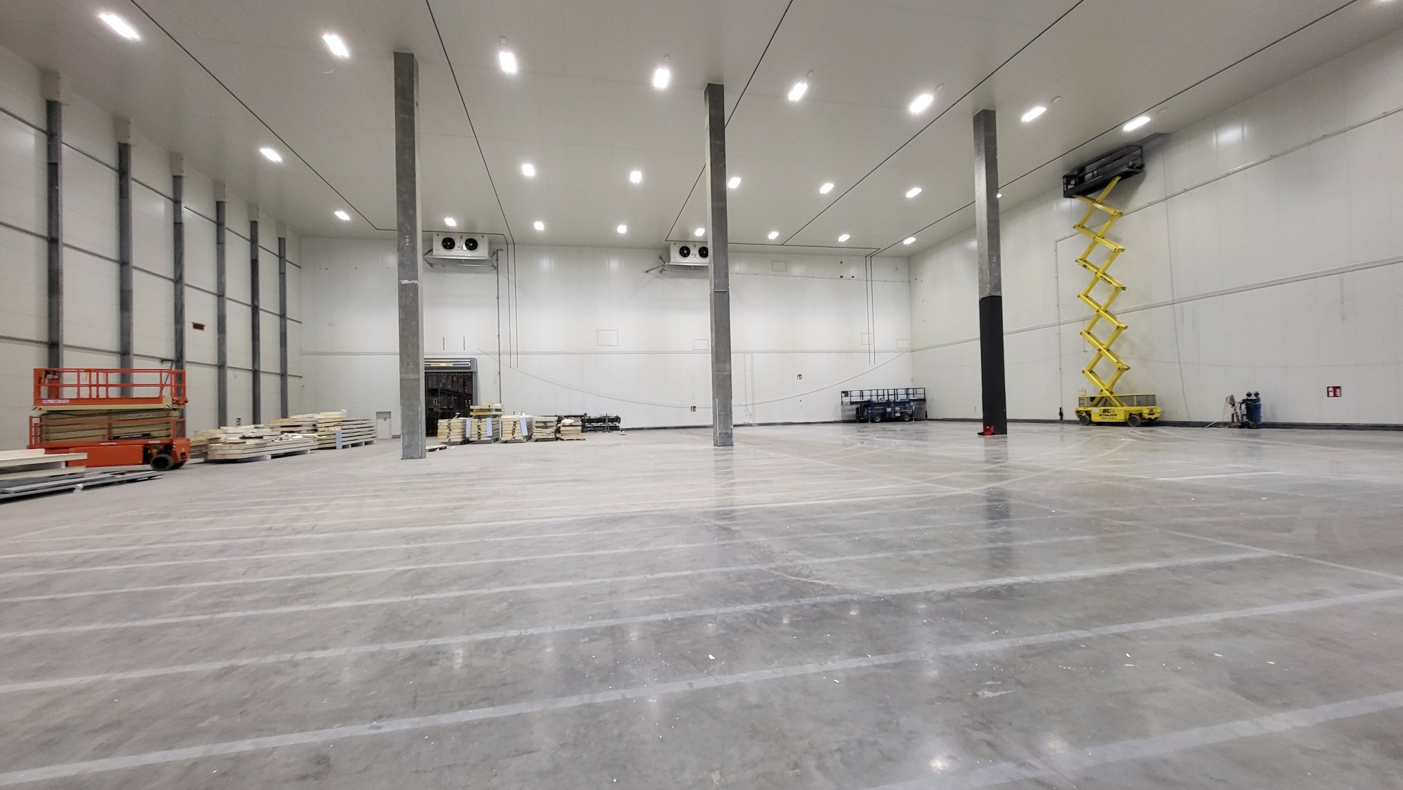 Wnętrze hali magazynowej w trakcie montażu regałów wysokiego składowania; widoczne podnośniki nożycowe, materiały budowlane i betonowa posadzka z wyznaczonymi liniami.