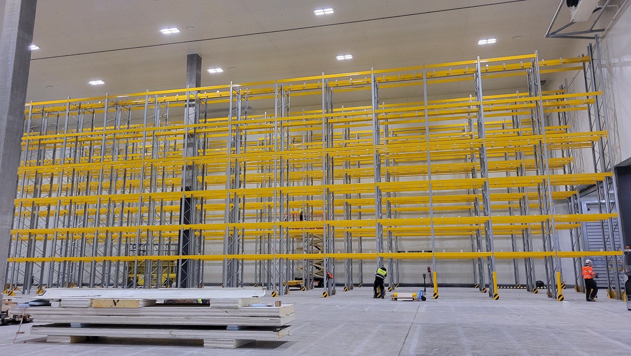 Montaż regałów magazynowych wysokiego składowania w centrum dystrybucyjnym LIDL, widoczni pracownicy w odblaskowych kamizelkach, podnośnik nożycowy, elementy konstrukcyjne na paletach, surowa...