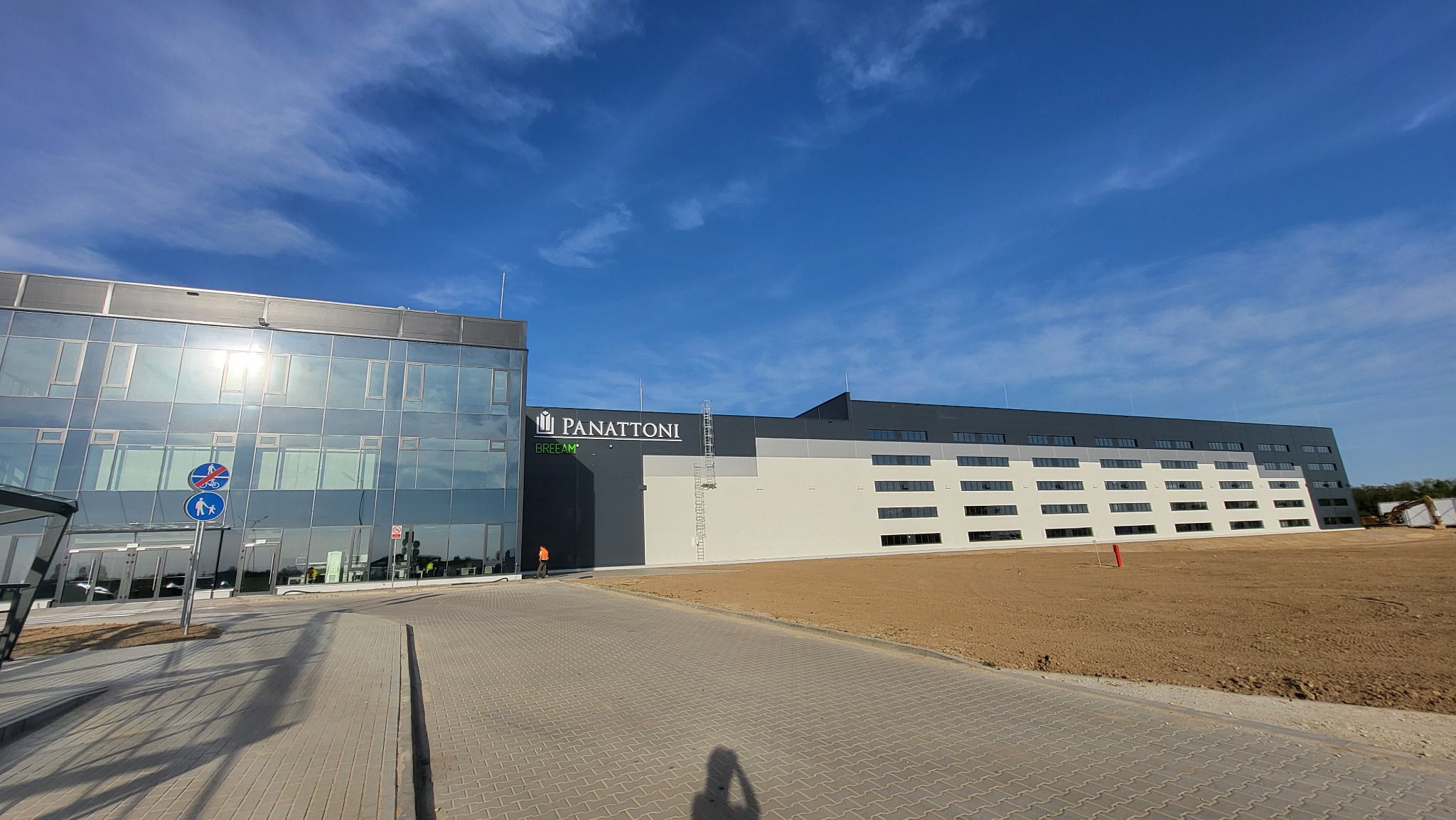 Rozległy widok na nowoczesny budynek magazynowy Panattoni w Sulechowie, z częścią biurową o szklanej fasadzie, placem budowy w tle i osobą w pomarańczowej kamizelce, sugerującą nadzór robót.