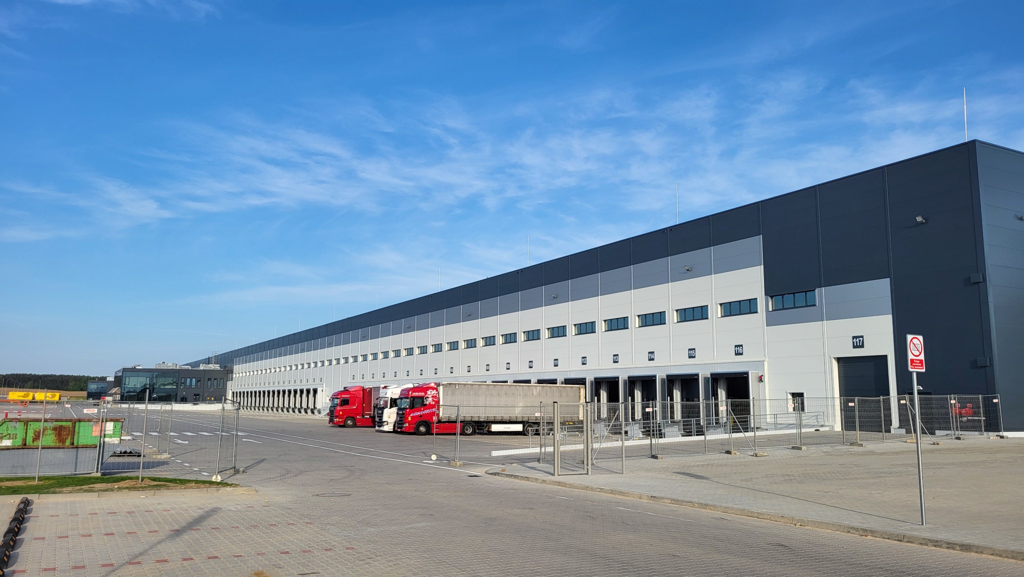 Nowoczesna hala magazynowa Panattoni Sulechów z rampami załadunkowymi i zaparkowanymi czerwonymi ciężarówkami w słoneczny dzień.
