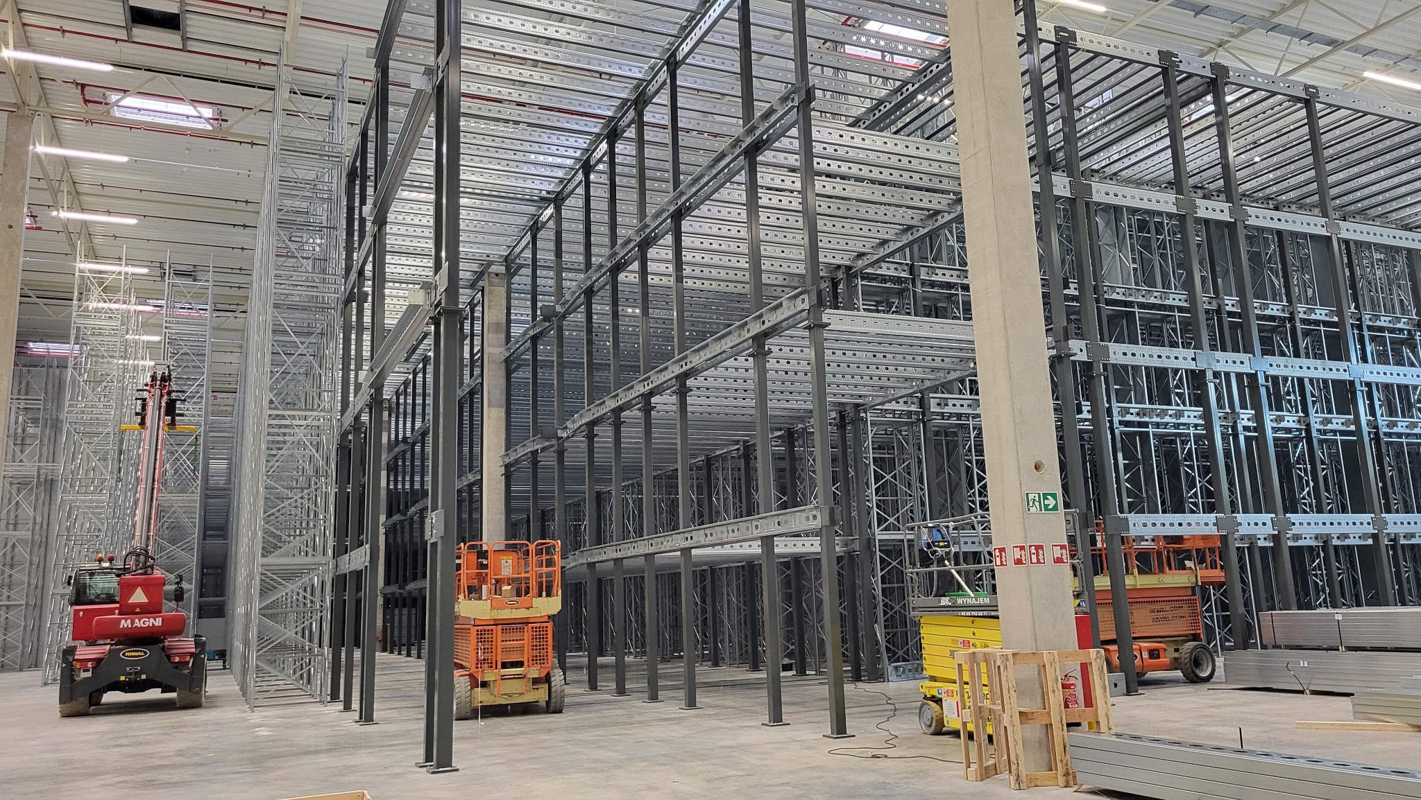 Wnętrze hali magazynowej w trakcie montażu konstrukcji stalowych regałów, widoczne podnośniki koszowe i elementy konstrukcyjne.