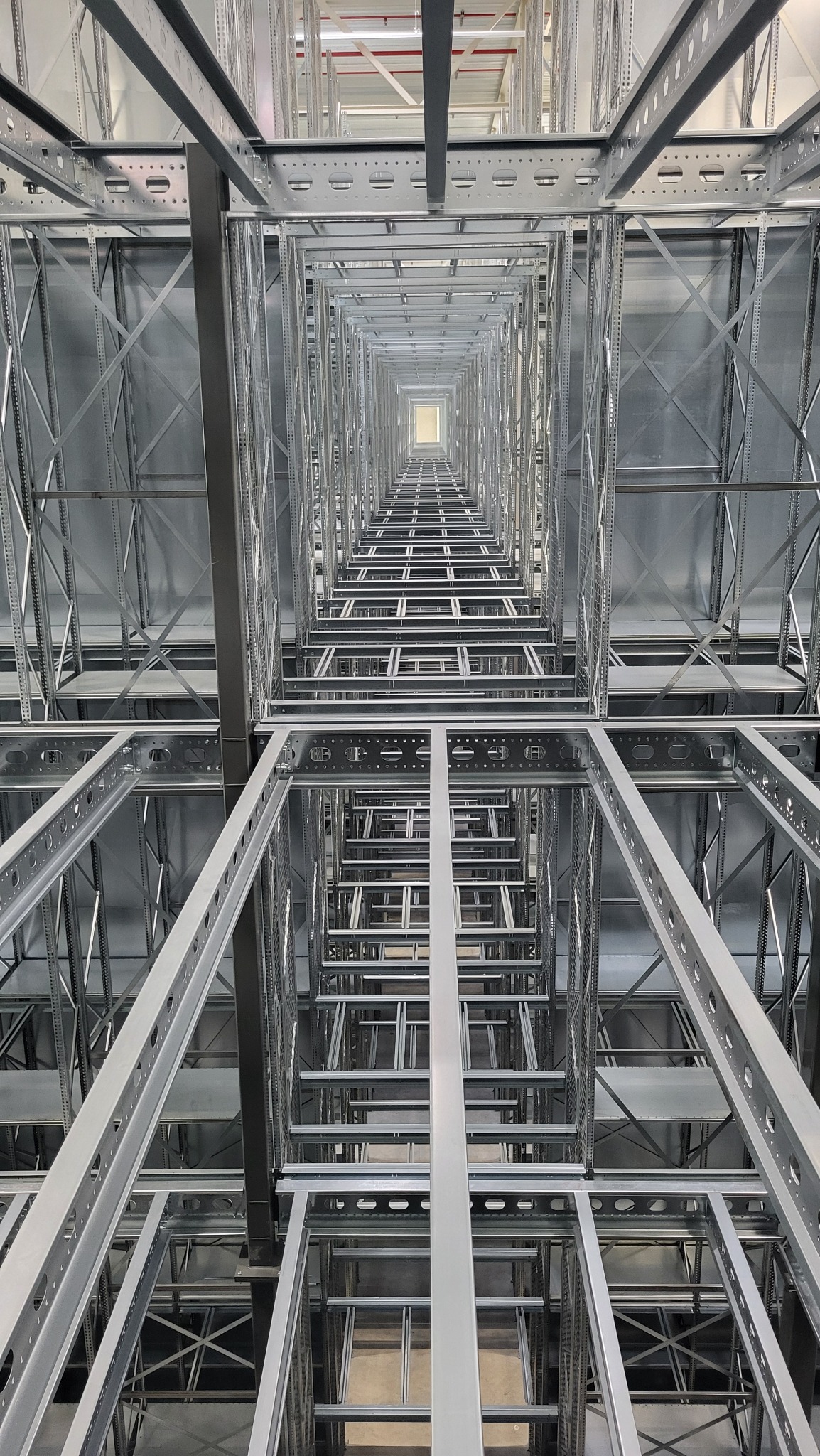 Wnętrze hali magazynowej w trakcie montażu konstrukcji stalowej, widok z dołu wzdłuż pionowych słupów, perspektywa zbieżna ku górze.