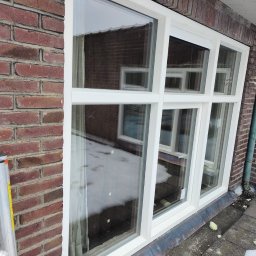 MK Fenster - Maciej Kaszczyk @oknamaciek - Białe okno PCV z podziałami, zamontowane w ścianie z czerwonej cegły, widoczne odbicie budynku i resztki śniegu na parapecie.