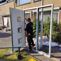 MK Fenster - Maciej Kaszczyk @oknamaciek - Montaż drzwi wejściowych z trzema małymi okienkami; widoczny instalator w czarnym uniformie, żółta skrzynka narzędziowa na trawie oraz rama drzwiowa oparta o słup.