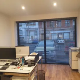 Biuro z widokiem na ulicę przez poziome listwy żaluzji; na pierwszym planie biurko z dwoma monitorami i stertą dokumentów.