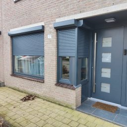 MK Fenster - Maciej Kaszczyk @oknamaciek - Szara elewacja domu z dwoma oknami z częściowo opuszczonymi roletami zewnętrznymi i nowoczesnymi drzwiami wejściowymi z numerem 17.