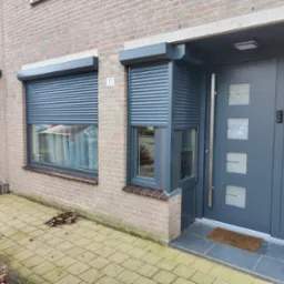 Szara elewacja domu z dwoma oknami z częściowo opuszczonymi roletami zewnętrznymi i nowoczesnymi drzwiami wejściowymi z numerem 17.