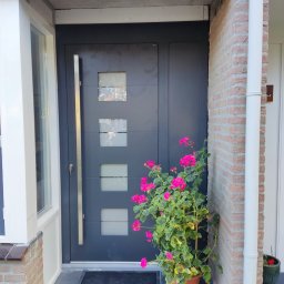 MK Fenster - Maciej Kaszczyk @oknamaciek - Nowoczesne, antracytowe drzwi wejściowe z czterema kwadratowymi przeszkleniami, ozdobione doniczką z kwitnącymi na różowo pelargoniami, widoczne z zewnątrz budynku.