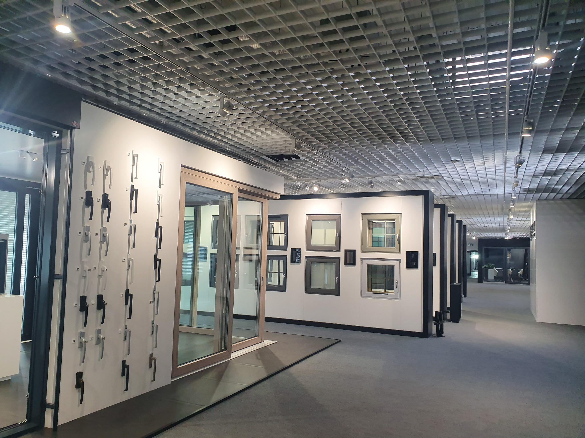 Ekspozycja okien i drzwi w salonie: przesuwne drzwi tarasowe z drewnianą ramą, różne modele okien na ścianie oraz kolekcja klamek na białym tle, oświetlenie sufitowe.