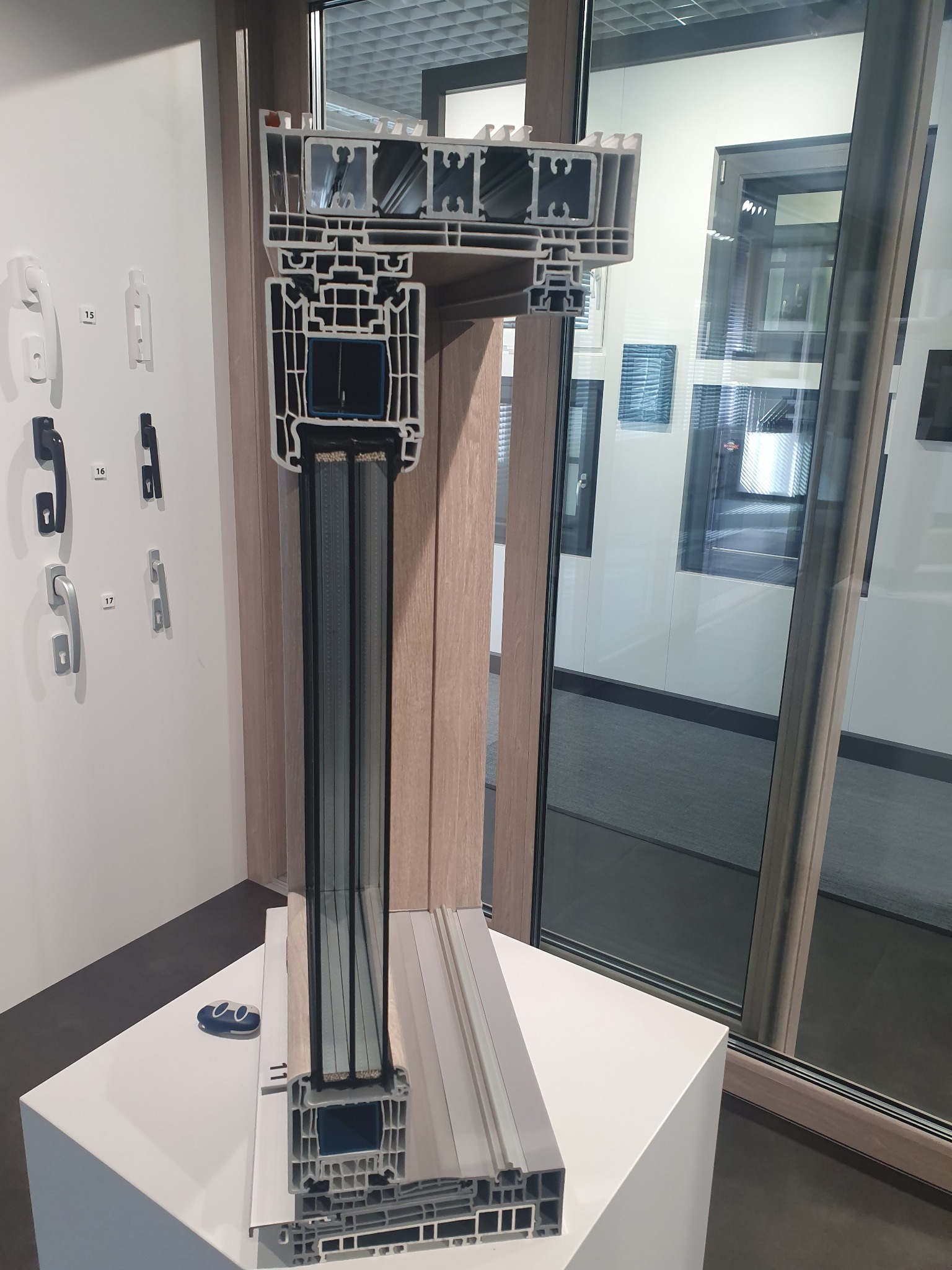 Przekrój okna z widoczną wielokomorową konstrukcją ramy i szyb zespolonych, prezentowany na białym postumencie obok ekspozycji klamek i gotowych okien