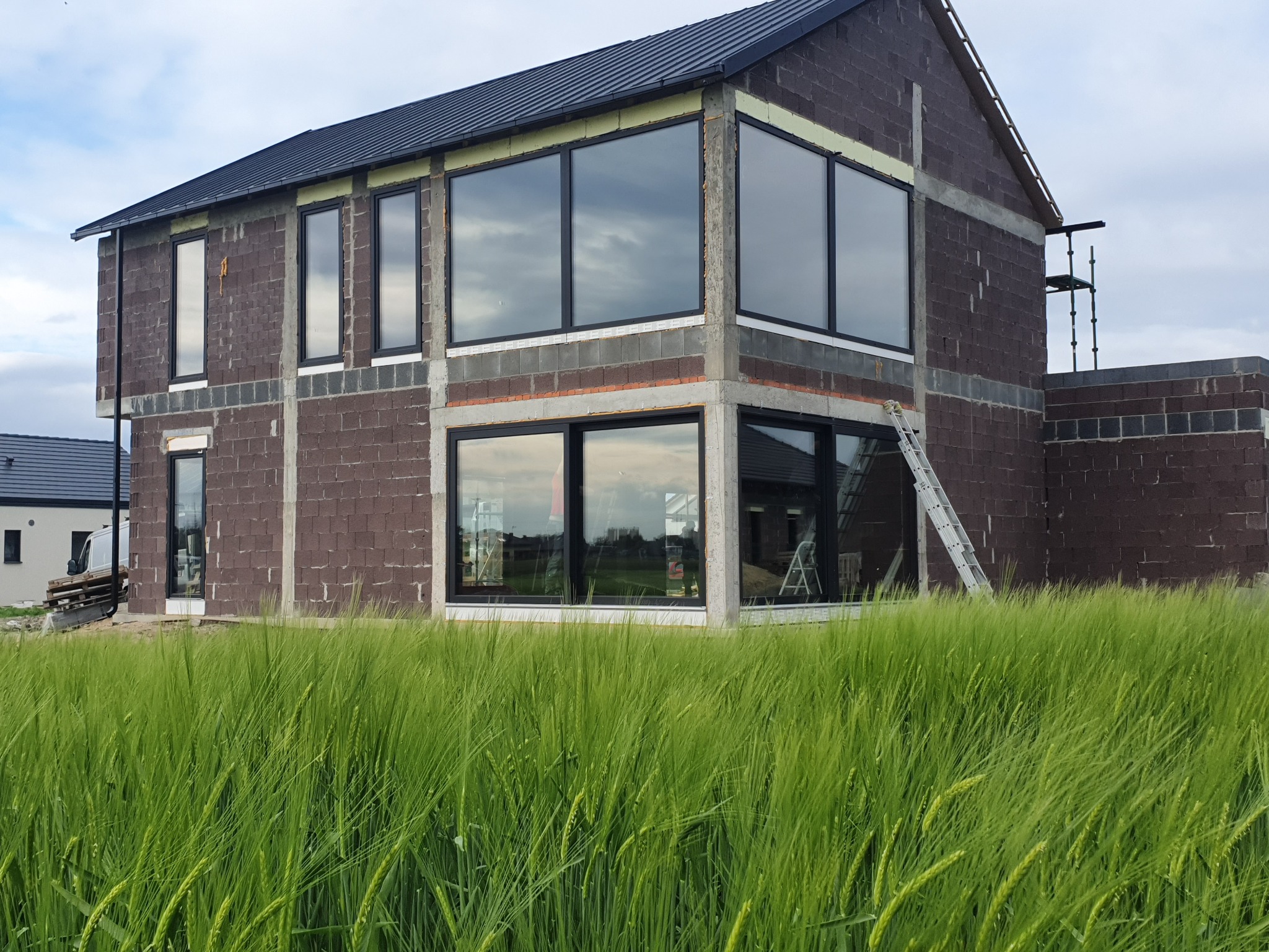 Surowy budynek w trakcie budowy z zamontowanymi dużymi, czarnymi oknami, widoczny przez łan zielonego zboża, z rusztowaniem z boku.