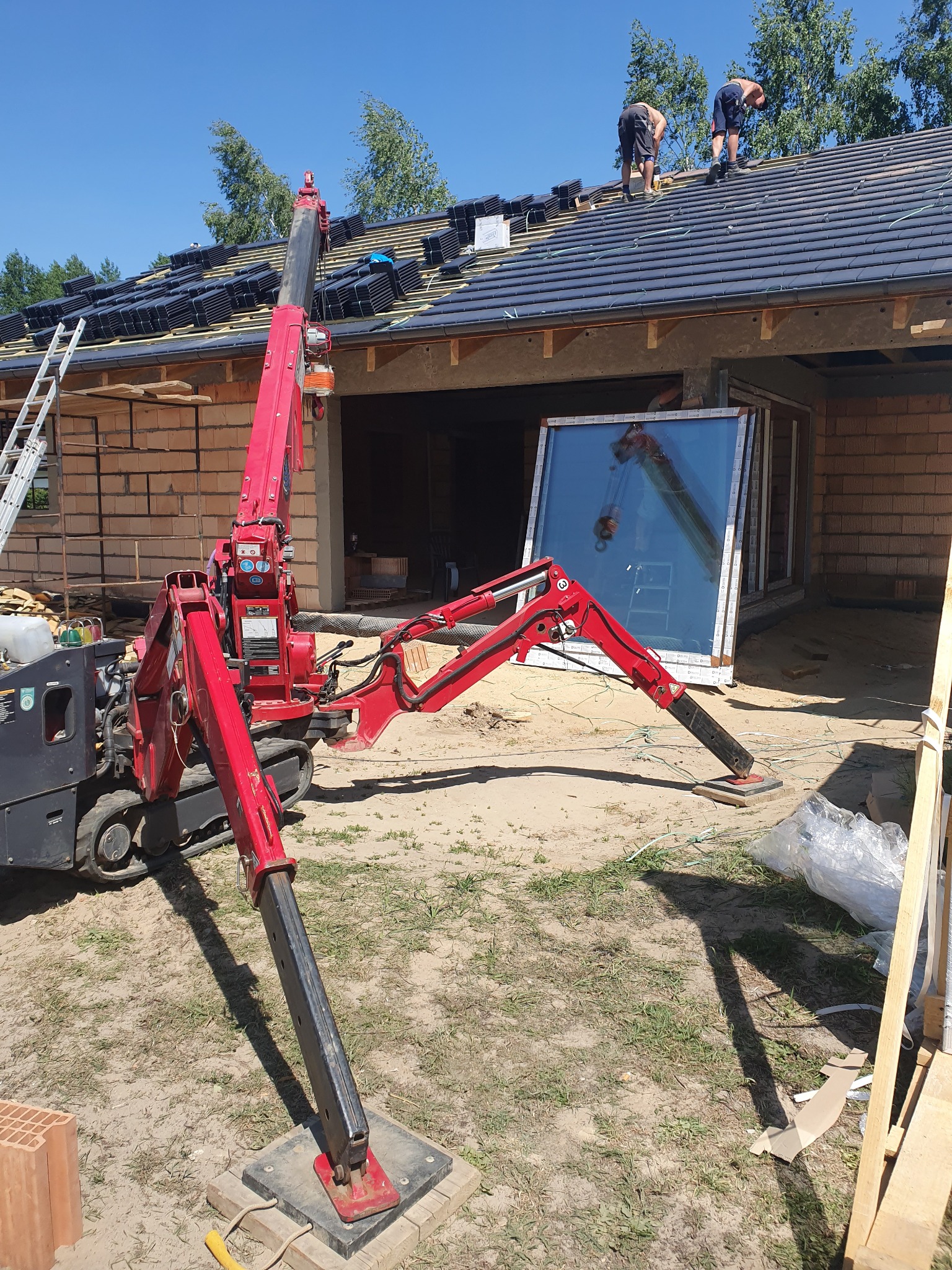 Montaż okien PCV w nowym domu z wykorzystaniem mini żurawia, widoczni dekarze układający dachówkę