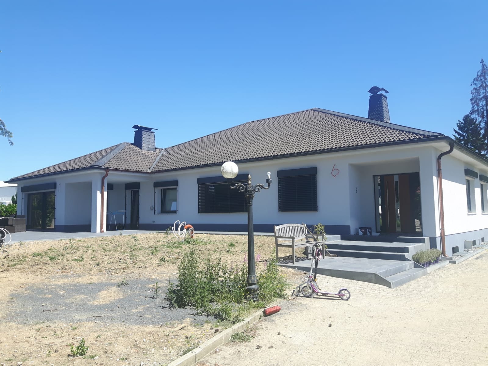 Parterowy dom jednorodzinny z ciemnymi żaluzjami zewnętrznymi, widok z przodu na ogród z lampą i hulajnogą.