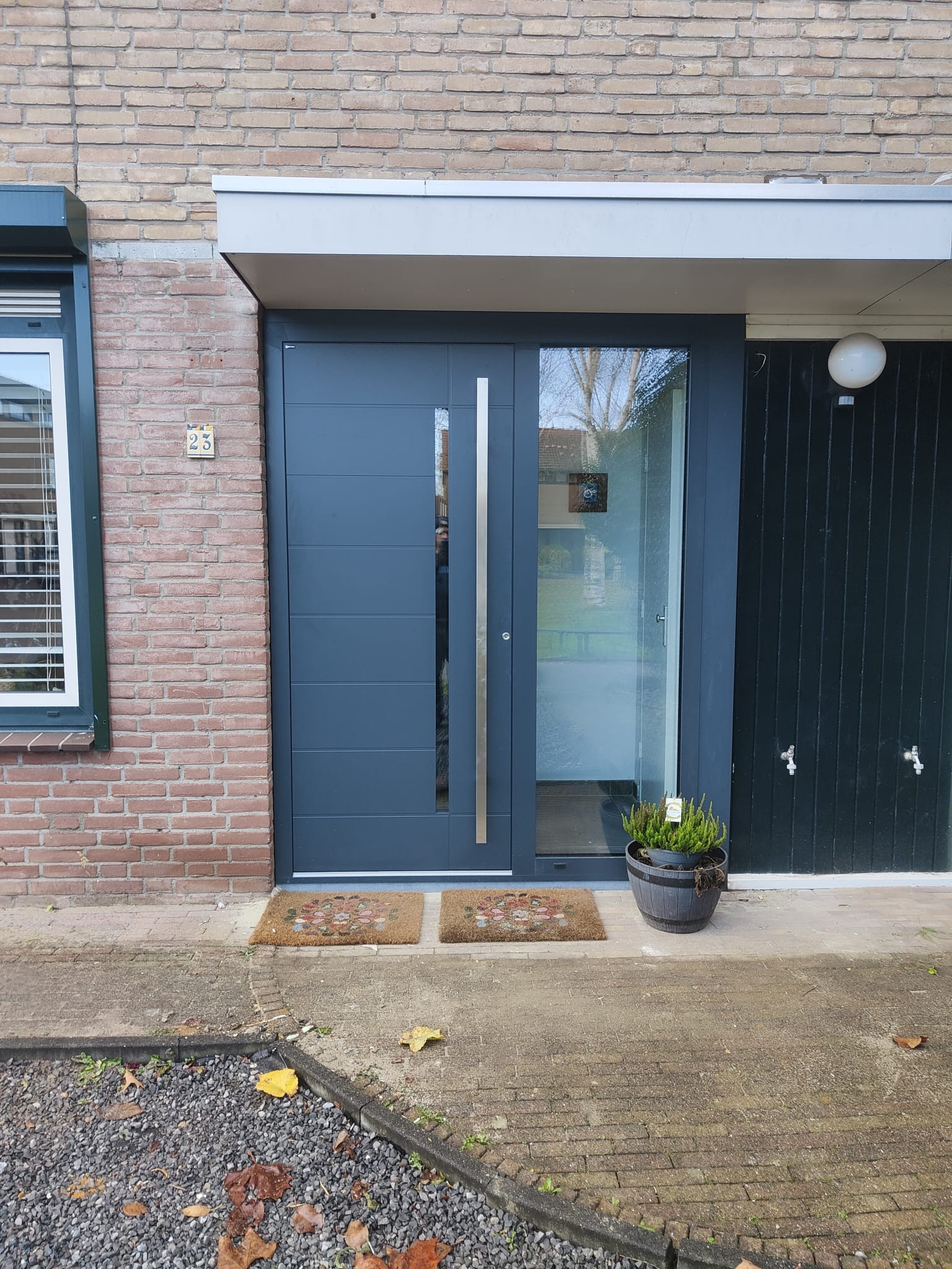 Nowoczesne drzwi wejściowe w kolorze grafitowym z pionowym przeszkleniem, ozdobione dwoma wycieraczkami i doniczką z rośliną, na tle ceglanej ściany i ciemnej, drewnianej zabudowy.