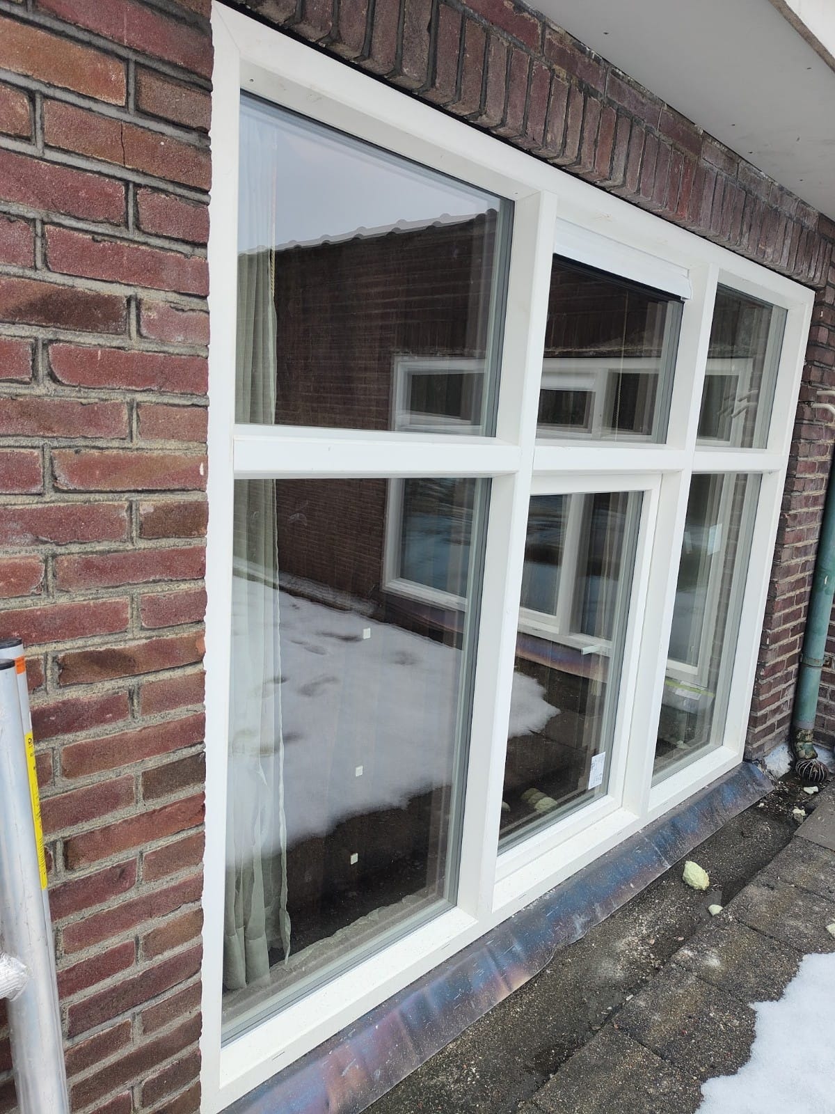 Białe okno PCV z podziałami, zamontowane w ścianie z czerwonej cegły, widoczne odbicie budynku i resztki śniegu na parapecie.