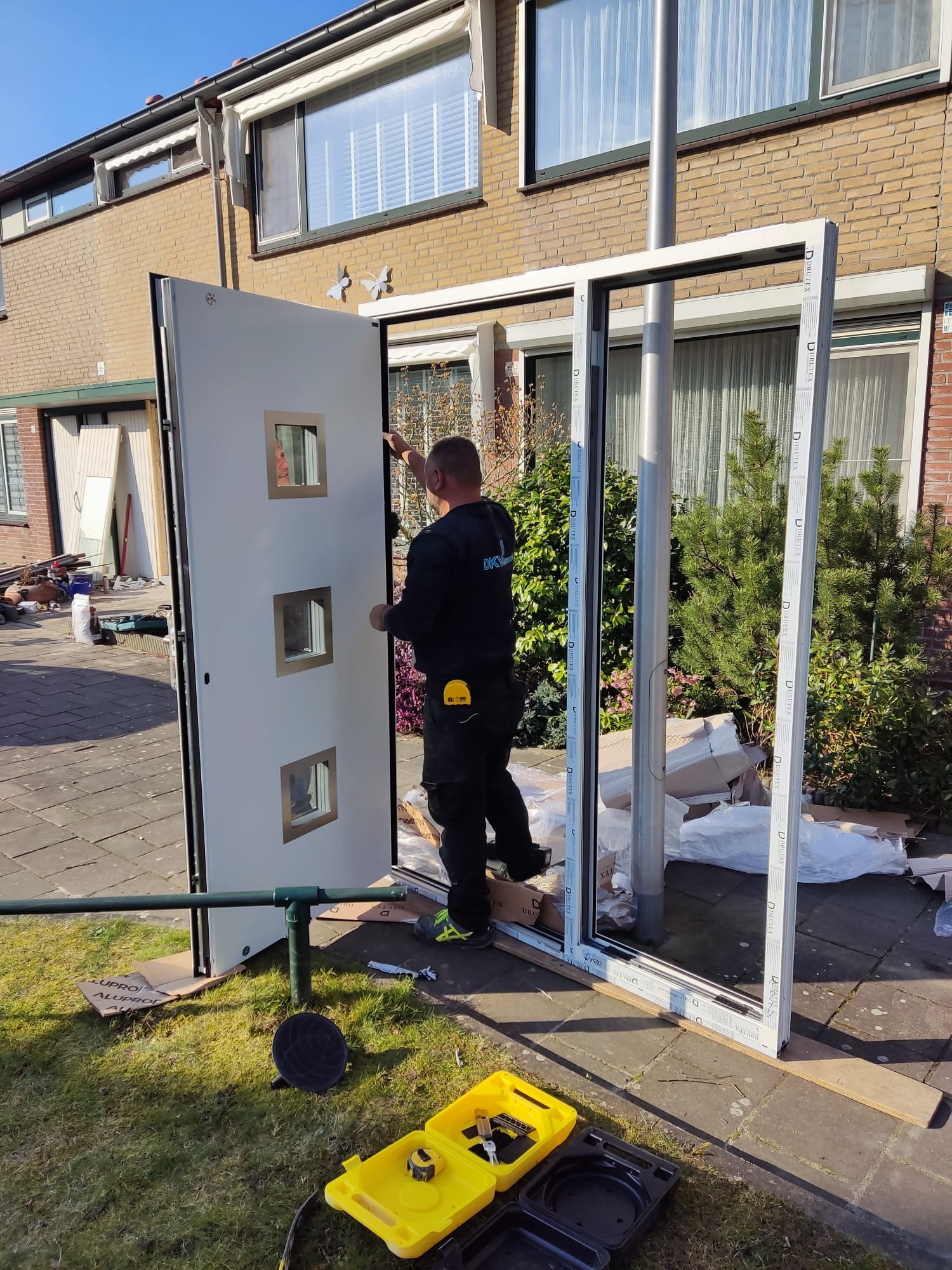 Montaż drzwi wejściowych z trzema małymi okienkami; widoczny instalator w czarnym uniformie, żółta skrzynka narzędziowa na trawie oraz rama drzwiowa oparta o słup.