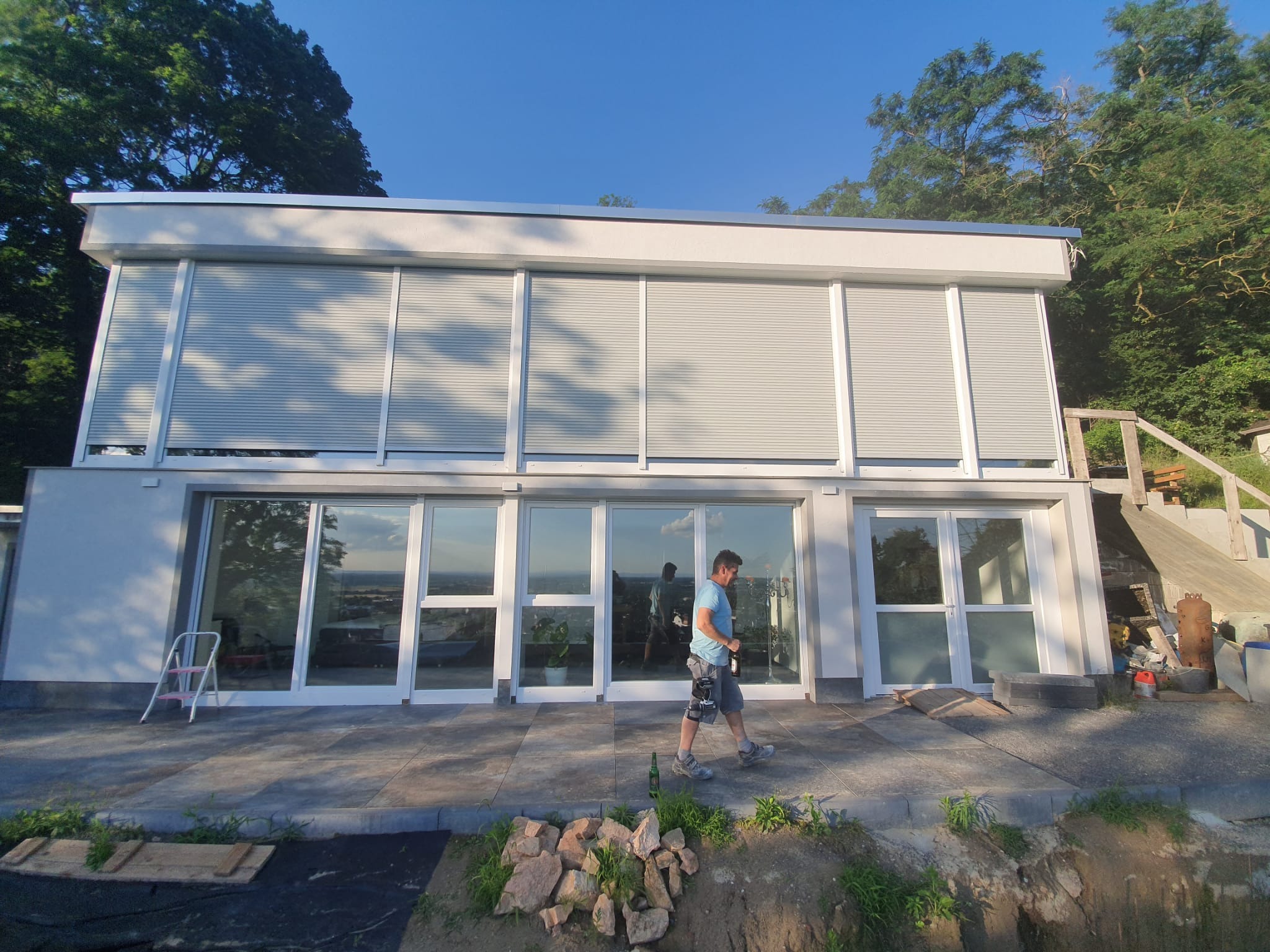 Nowoczesny budynek z białymi ramami okiennymi i szarymi żaluzjami fasadowymi, mężczyzna przechodzi przed budynkiem, na pierwszym planie teren w trakcie prac ziemnych.