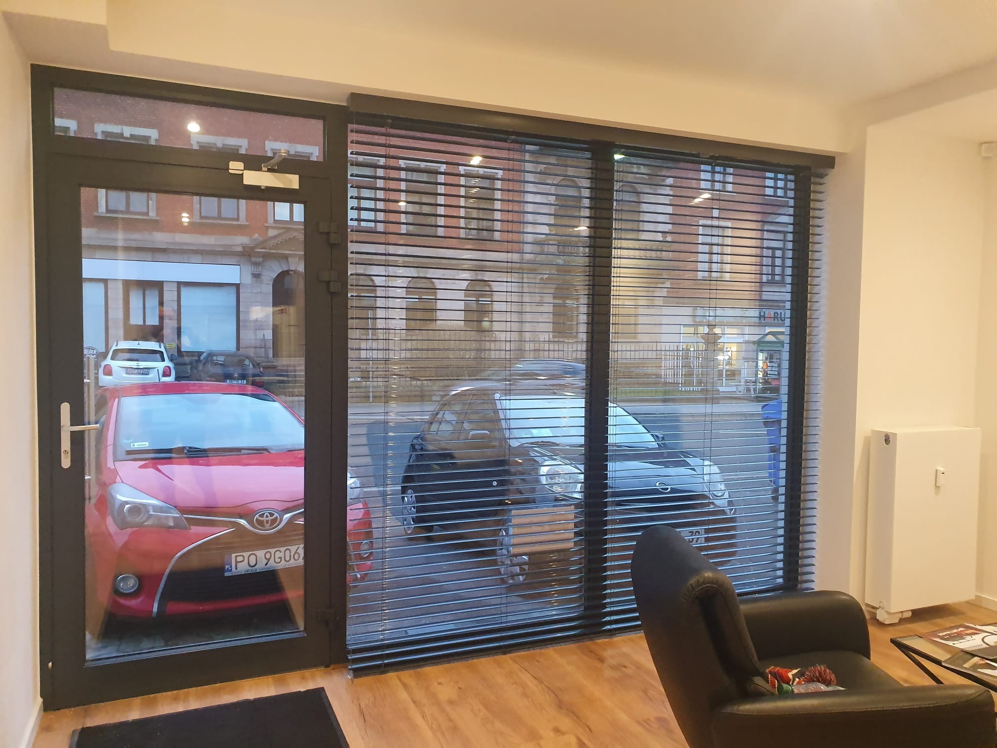 Wnętrze pomieszczenia z dużym oknem i drzwiami balkonowymi, osłoniętymi ciemnymi żaluzjami poziomymi, widok na ulicę z zaparkowanymi samochodami, w pomieszczeniu fotel i grzejnik.