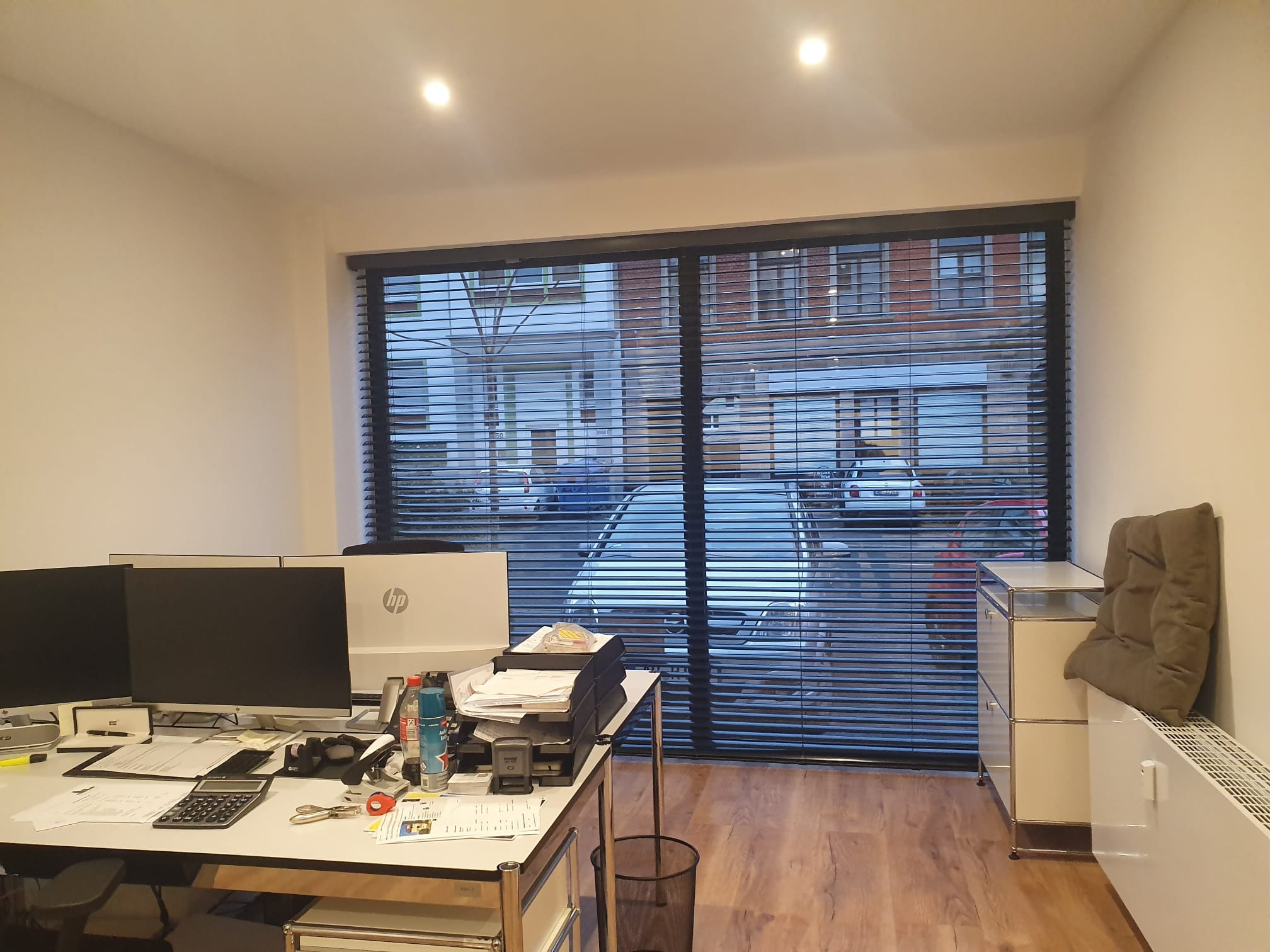 Biuro z widokiem na ulicę przez poziome listwy żaluzji; na pierwszym planie biurko z dwoma monitorami i stertą dokumentów.