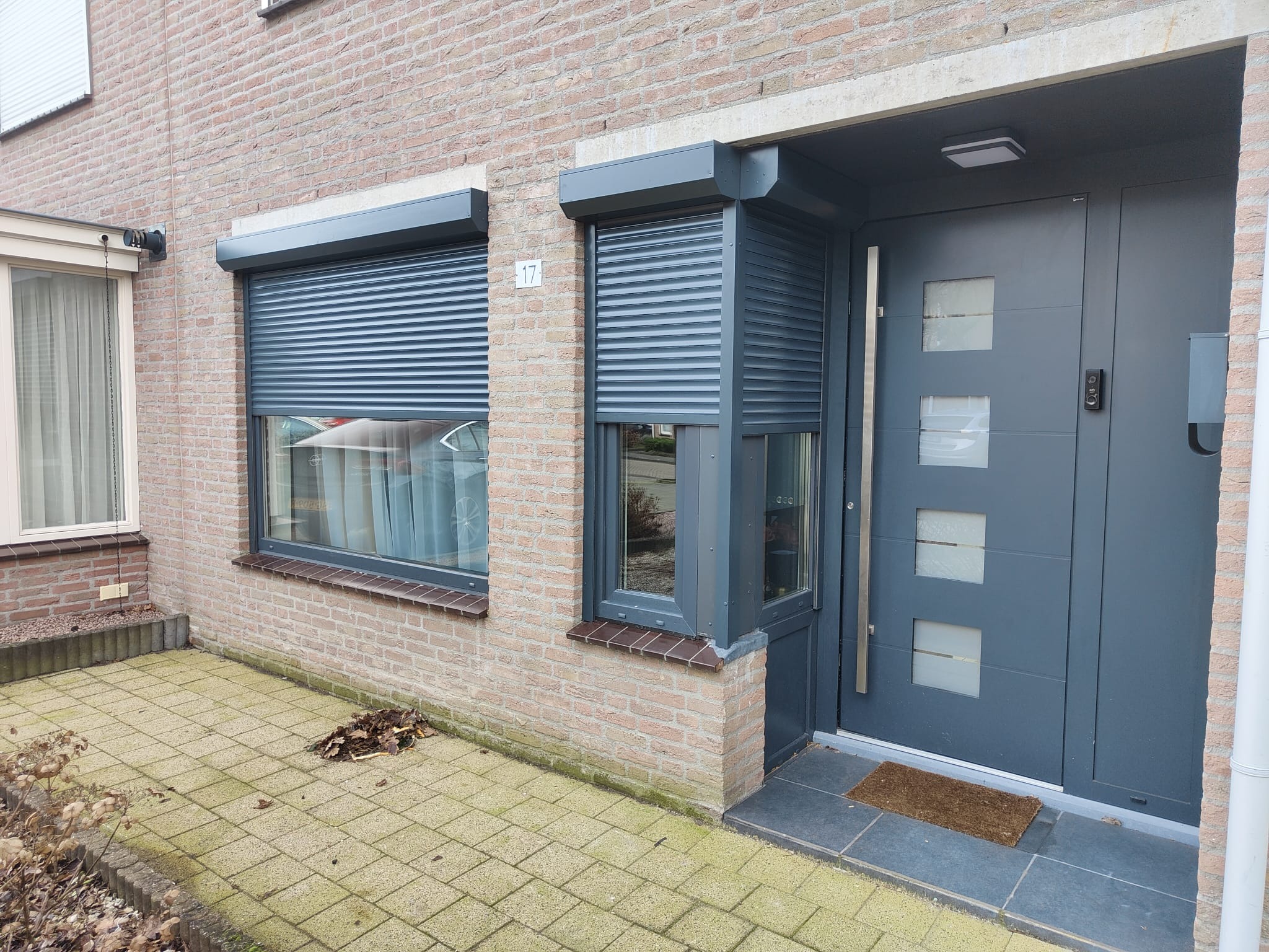 Szara elewacja domu z dwoma oknami z częściowo opuszczonymi roletami zewnętrznymi i nowoczesnymi drzwiami wejściowymi z numerem 17.