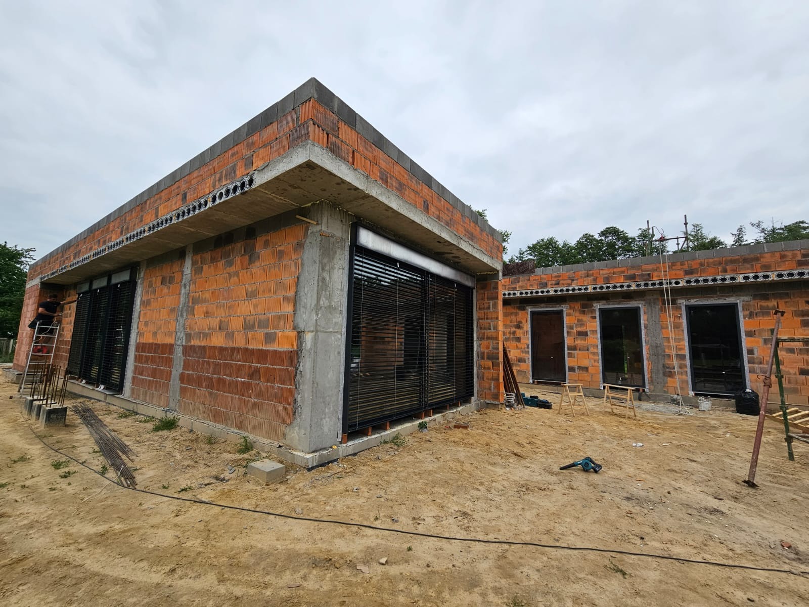 Montaż żaluzji fasadowych na surowej elewacji budynku z cegły, widoczne narzędzia i drabina.