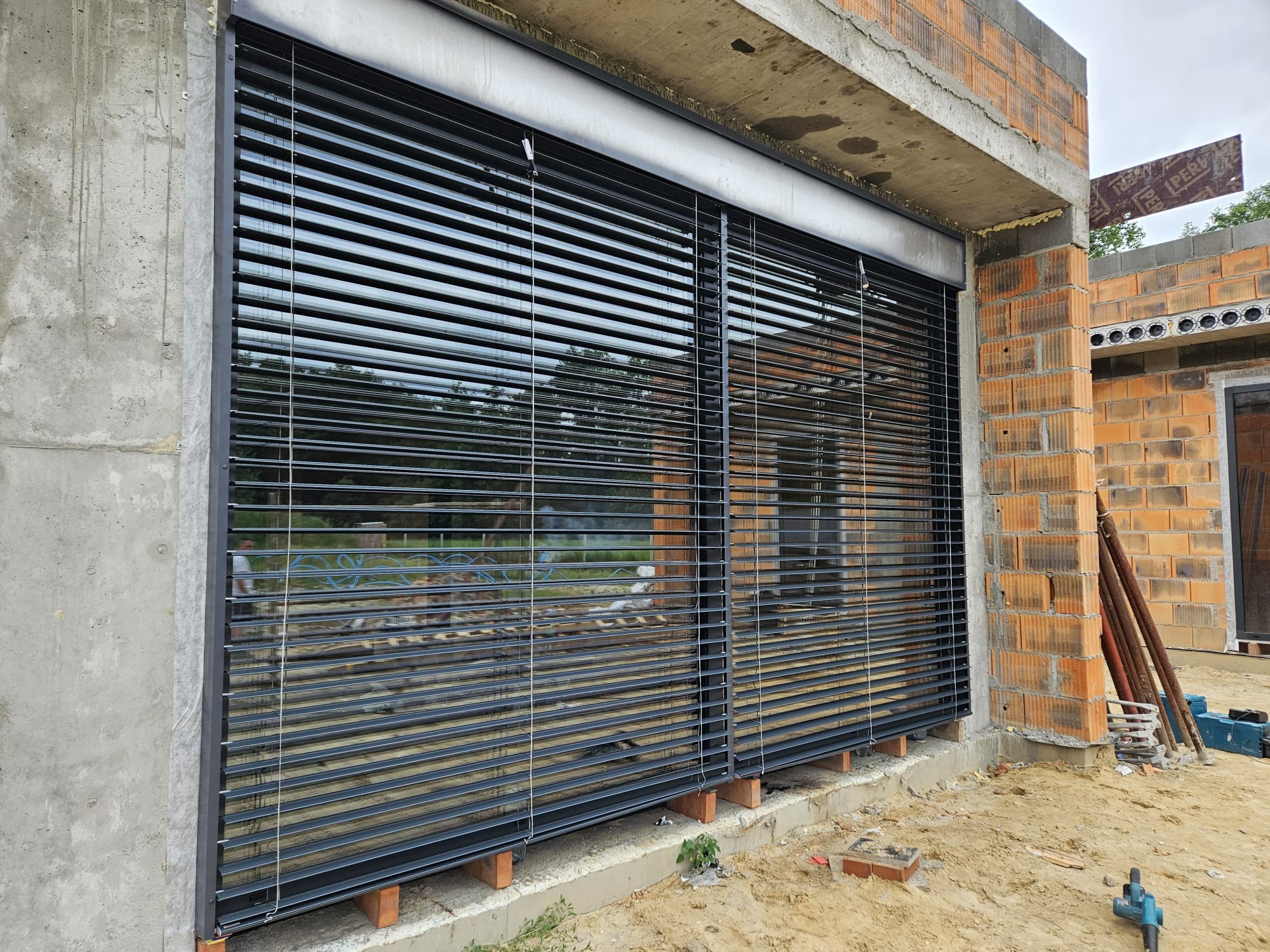 Zamontowane, czarne żaluzje fasadowe na surowym budynku z cegły, widoczne elementy konstrukcyjne i piasek.