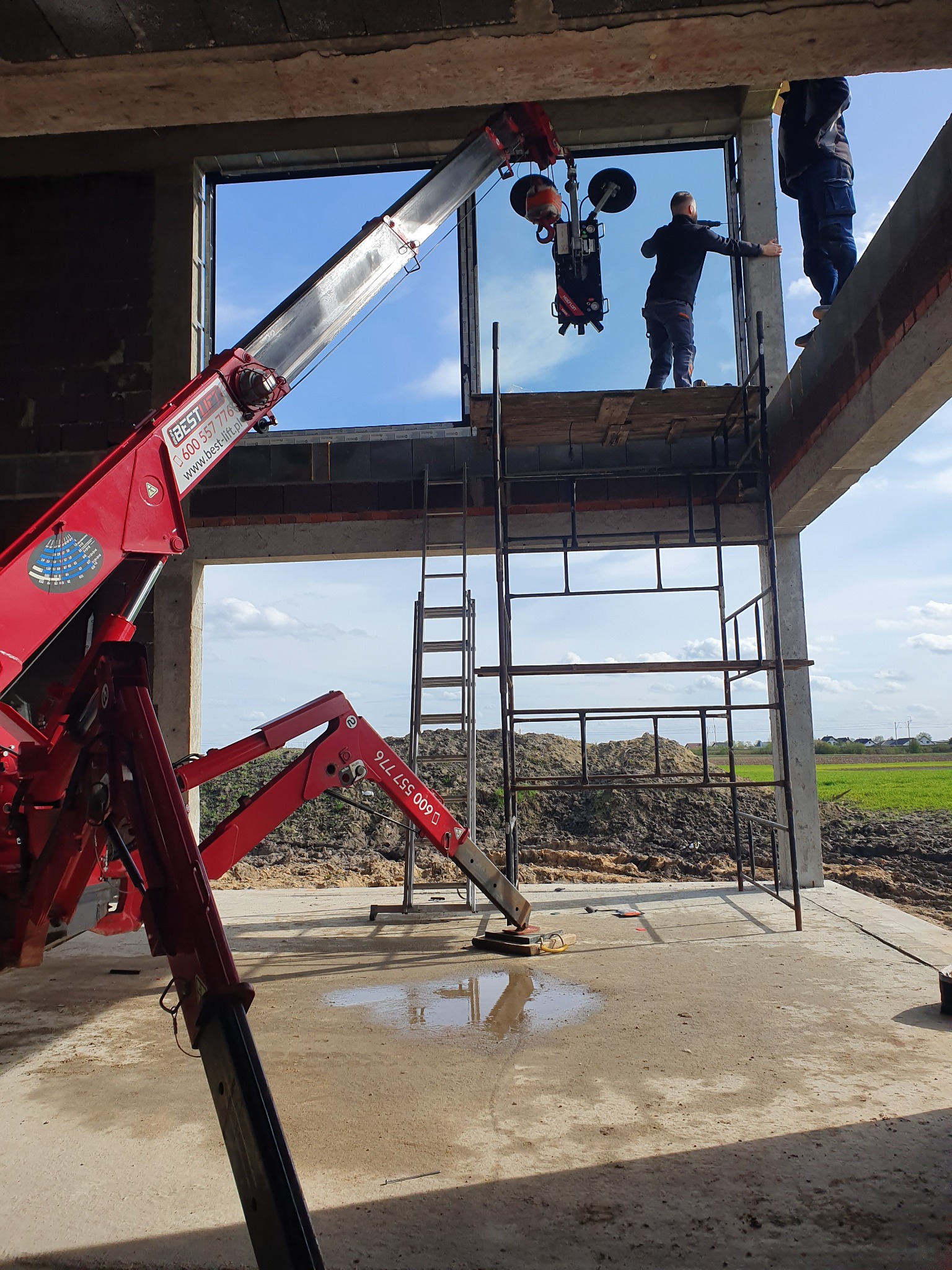 Montaż okna na budowie z użyciem przyssawki i dźwigu, widoczni dwaj monterzy na rusztowaniu, surowy stan wykończenia.