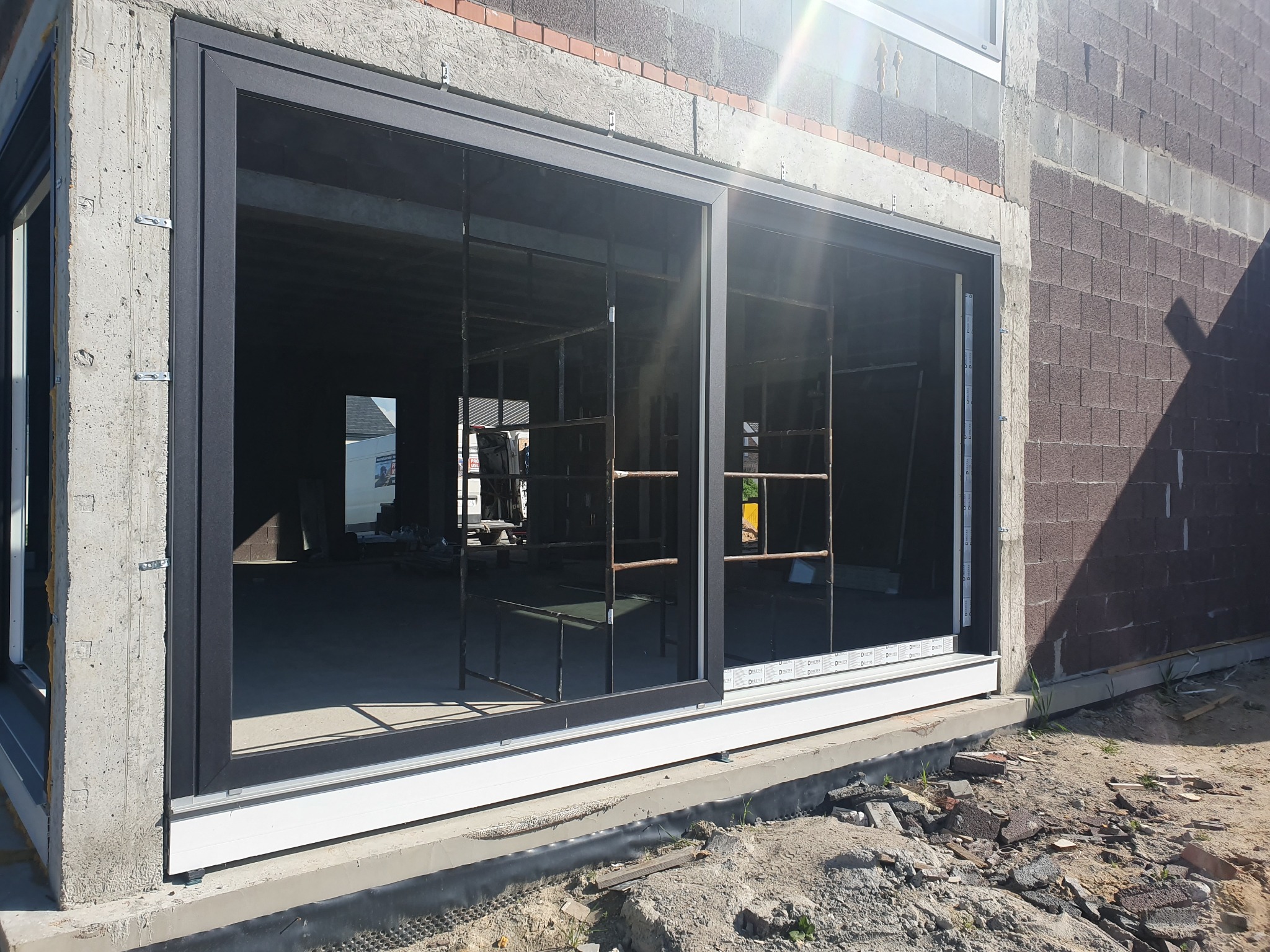 Montaż dużych przesuwnych okien z czarnymi ramami w surowym budynku, widoczne rusztowania wewnątrz.