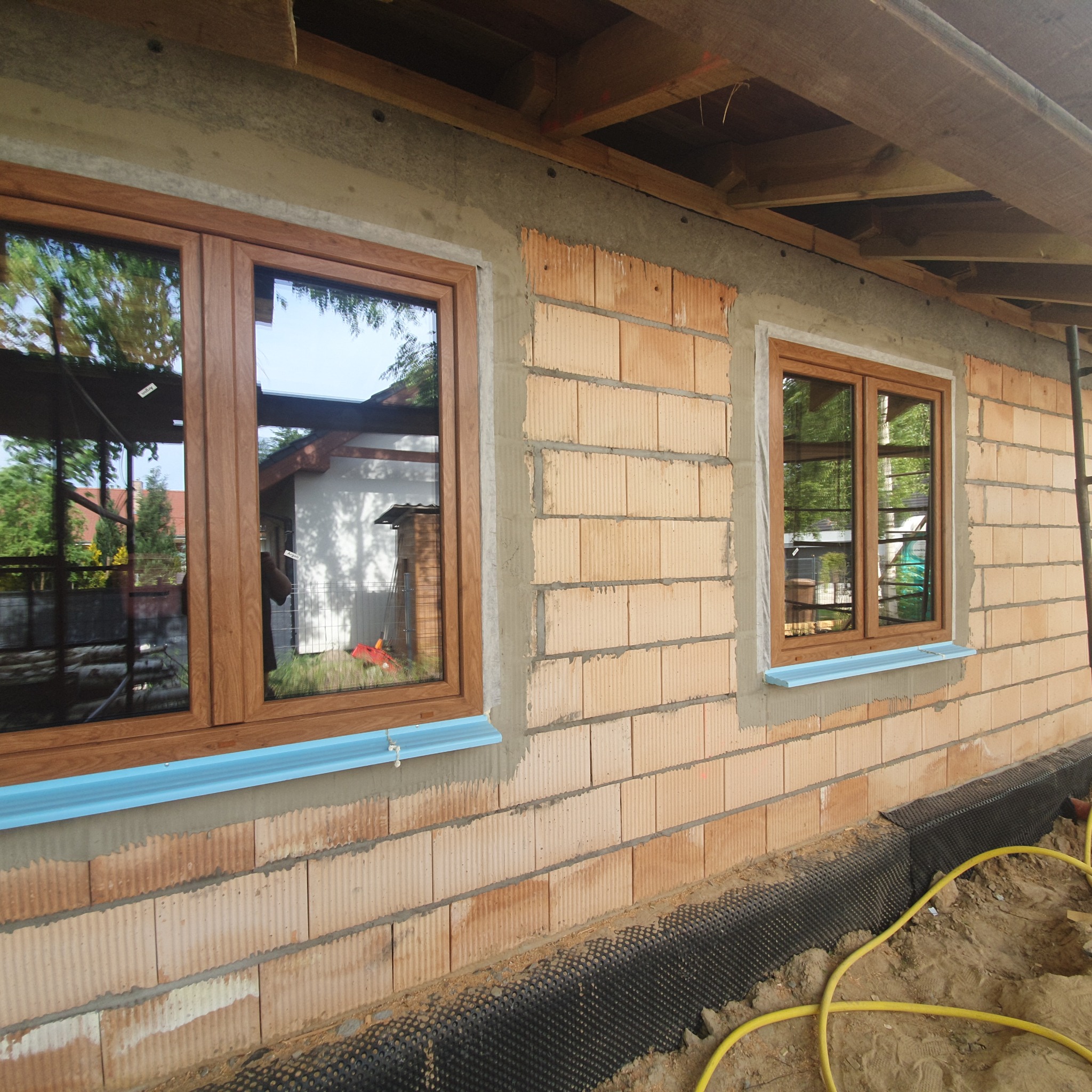 Montaż okien z brązowymi ramami w surowym murze z cegieł, widoczne zabezpieczenie parapetu pianką i folia fundamentowa przy ziemi, odzwierciedlenie drzew i budynku w szybach okiennych.