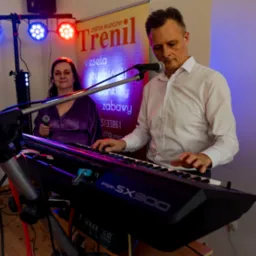 Muzyk grający na keyboardzie Yamaha PSR-SX900, obok wokalistka z mikrofonem, w tle oświetlenie sceniczne i baner reklamowy zespołu muzycznego.