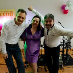 Trzy osoby, w tym kobieta w fioletowej sukience, pozują na tle banneru z nazwą 'Trenil' i sprzętu muzycznego, w tle balony.