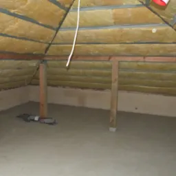Poddasze z izolacją z wełny mineralnej między krokwiami, widoczne drewniane słupy nośne i wylewka betonowa na podłodze, z resztkami zaprawy przy ścianie.