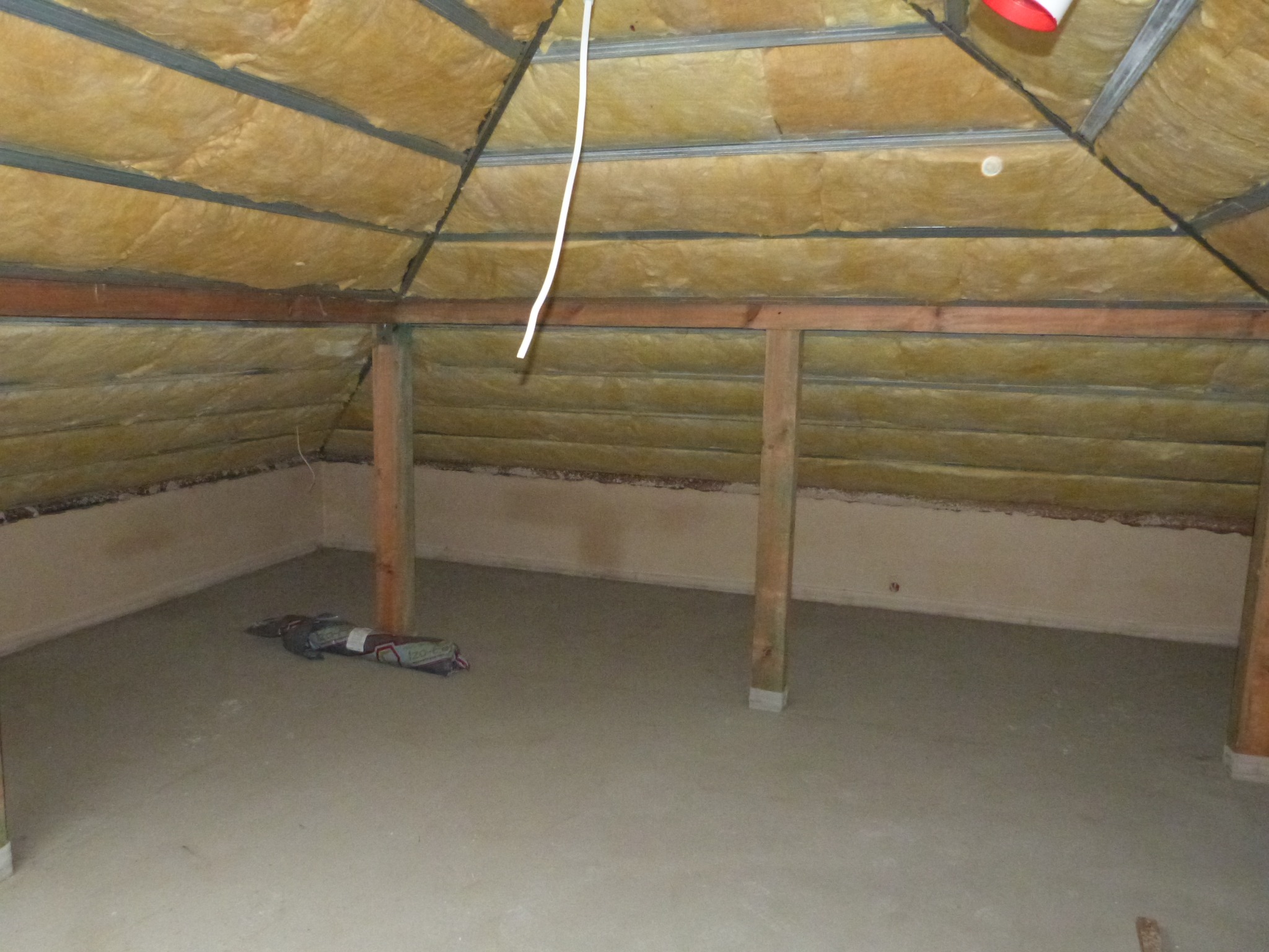 Poddasze z izolacją z wełny mineralnej między krokwiami, widoczne drewniane słupy nośne i wylewka betonowa na podłodze, z resztkami zaprawy przy ścianie.