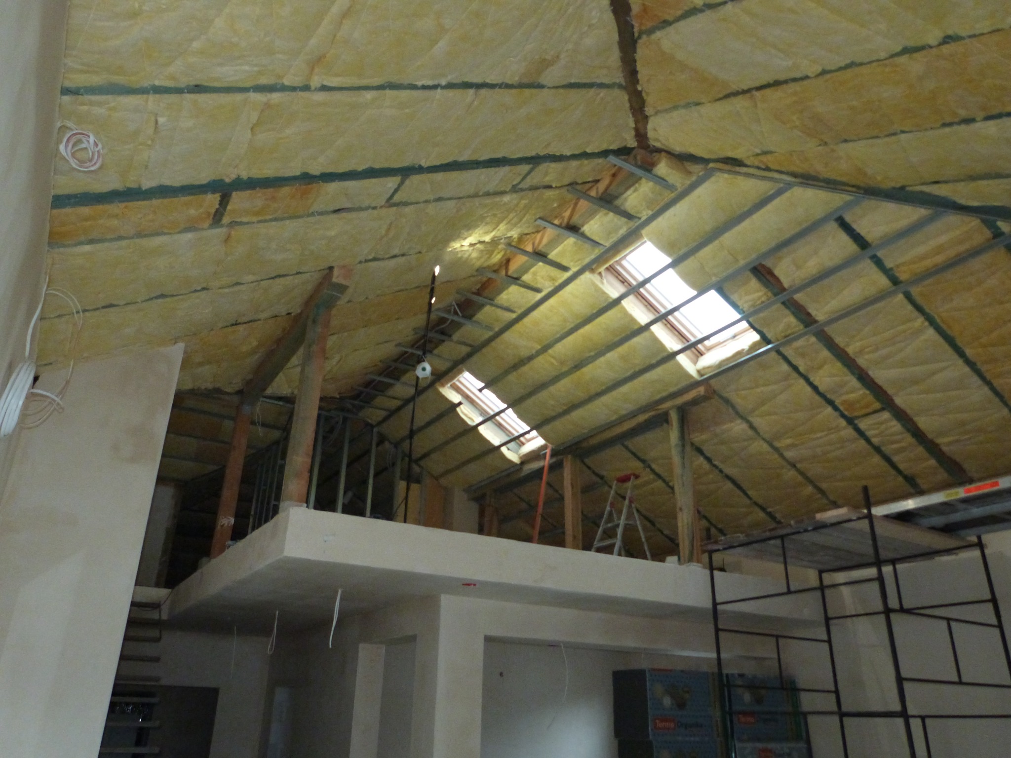 Poddasze w trakcie izolacji wełną mineralną, widoczne metalowe profile konstrukcyjne, okna dachowe i rusztowanie.