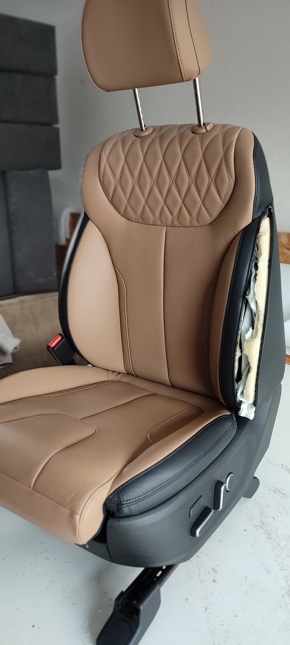 Fotel samochodowy z beżową skórzaną tapicerką, czarnymi akcentami i widocznym fragmentem wewnętrznej gąbki po stronie oparcia.