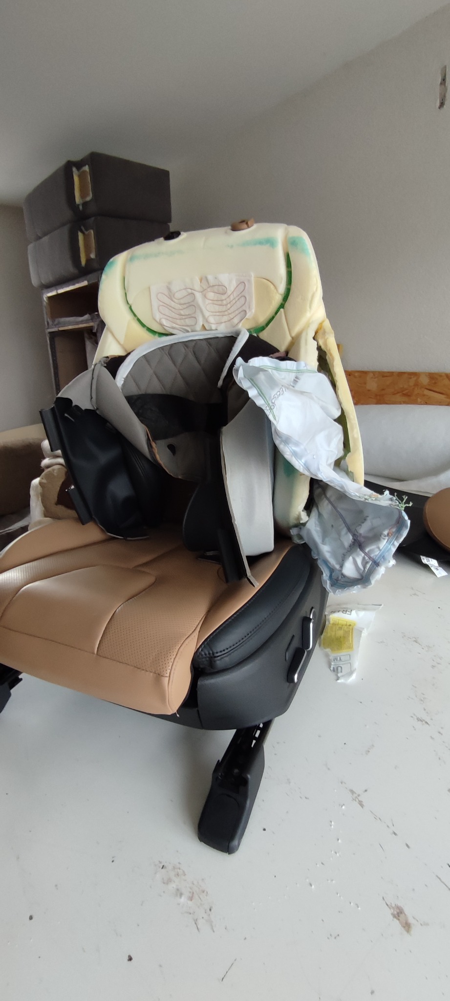 Rozłożone na części samochodowe siedzenie z beżową skórą i czarnymi plastikowymi elementami, widoczne warstwy gąbki i materiałów wyściełających, sugerujące proces naprawy lub renowacji.