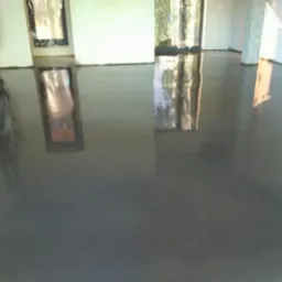 Świeżo wylana, gładka, szara posadzka w pustym pomieszczeniu z oknami, odbijająca światło, z widocznymi śladami pracy na ścianach.