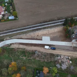 Widok z drona na nowo wybudowaną drogę dojazdową z betonu, łączącą istniejącą drogę z nowoczesnym domem w trakcie budowy, obok torów kolejowych i pola uprawnego.