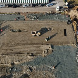 MURACZEWSKI PRACE ZIEMNE SPÓŁKA Z OGRANICZONĄ ODPOWIEDZIALNOŚCIĄ - Widok z góry na plac budowy z pracującymi maszynami: walcem, koparką i wywrotką. Część terenu wyrównana i utwardzona, widoczne ślady opon i stopy pracowników w pomarańczowych kamizelkach. W tle...