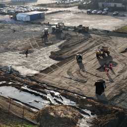 MURACZEWSKI PRACE ZIEMNE SPÓŁKA Z OGRANICZONĄ ODPOWIEDZIALNOŚCIĄ - Z góry widok placu budowy z ciężkim sprzętem, w tym walcami i koparkami, przygotowującego teren pod budowę, z widocznymi śladami opon i pracownikami w pomarańczowych kombinezonach.