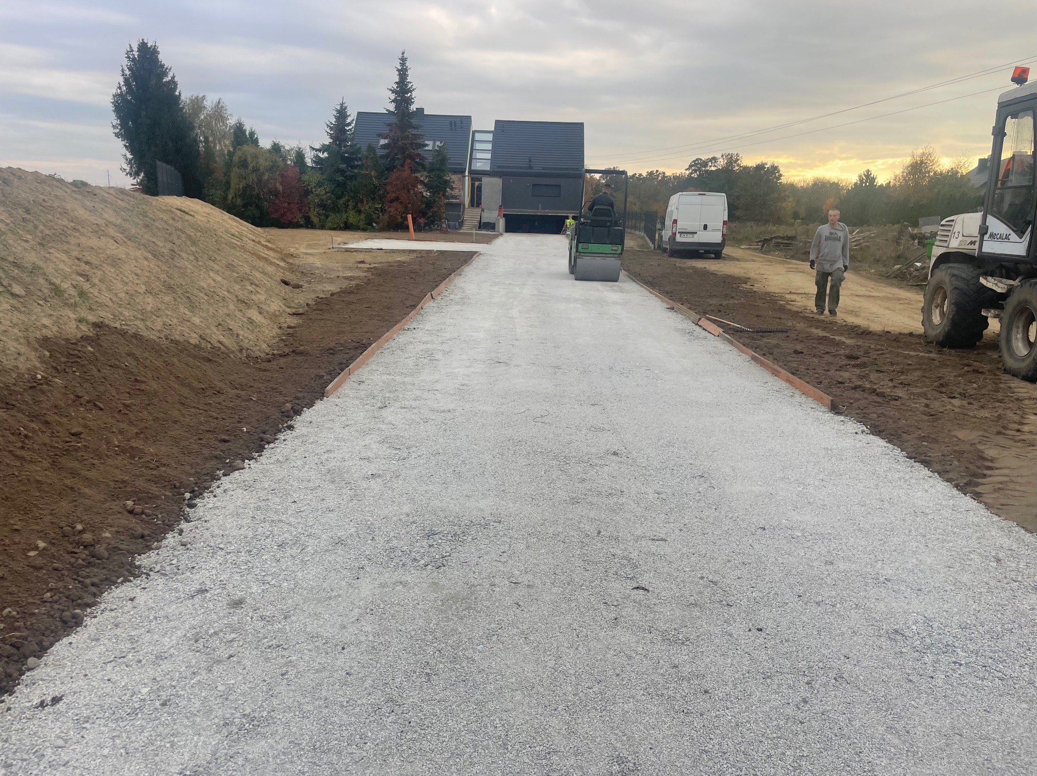 Walec drogowy ubija szarą nawierzchnię drogi dojazdowej do nowoczesnego domu z ciemną elewacją; widoczny pracownik w odblaskowej kamizelce, obok zaparkowany biały bus i koparko-ładowarka Mecalac.