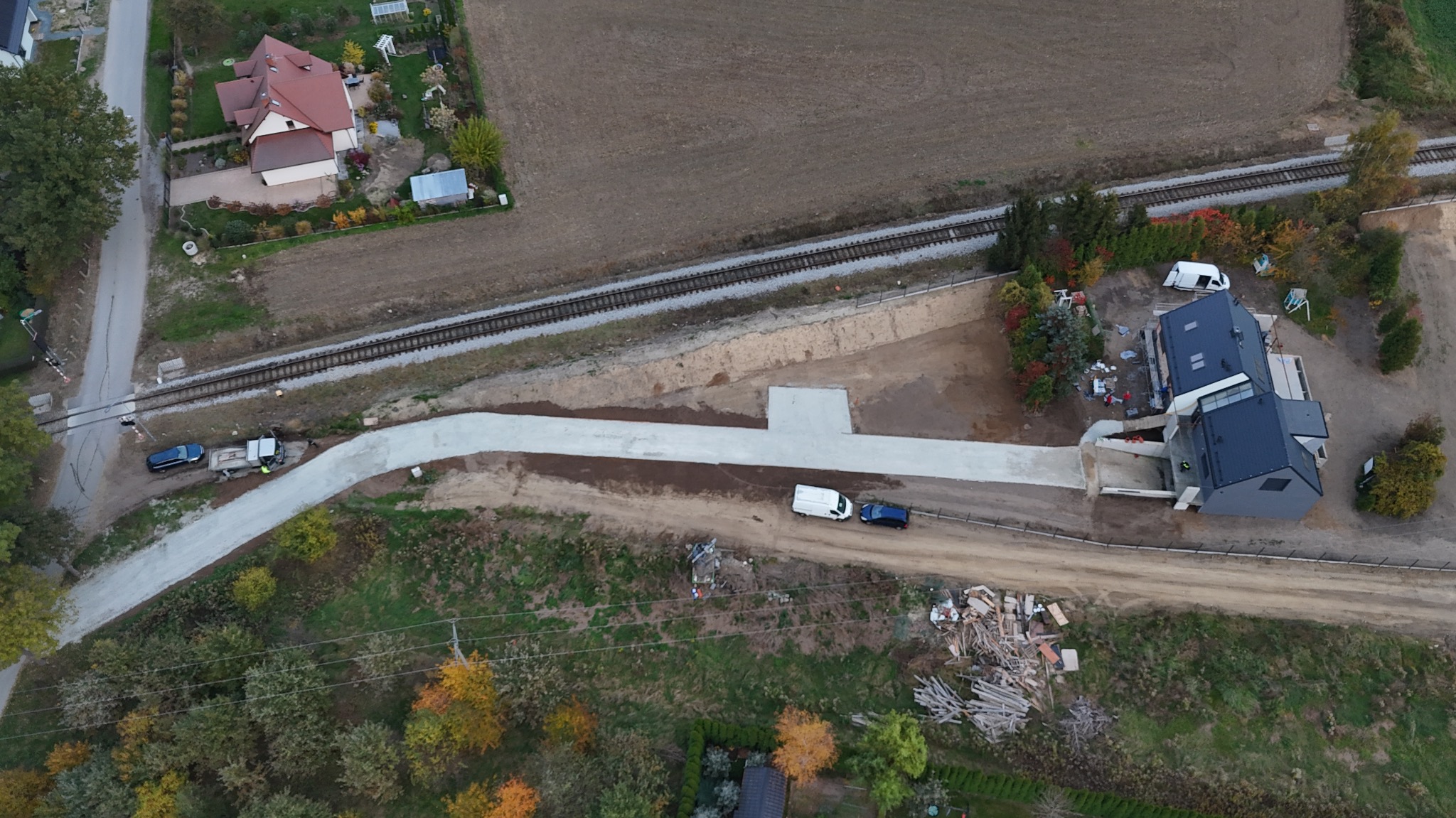 Widok z drona na nowo wybudowaną drogę dojazdową z betonu, łączącą istniejącą drogę z nowoczesnym domem w trakcie budowy, obok torów kolejowych i pola uprawnego.