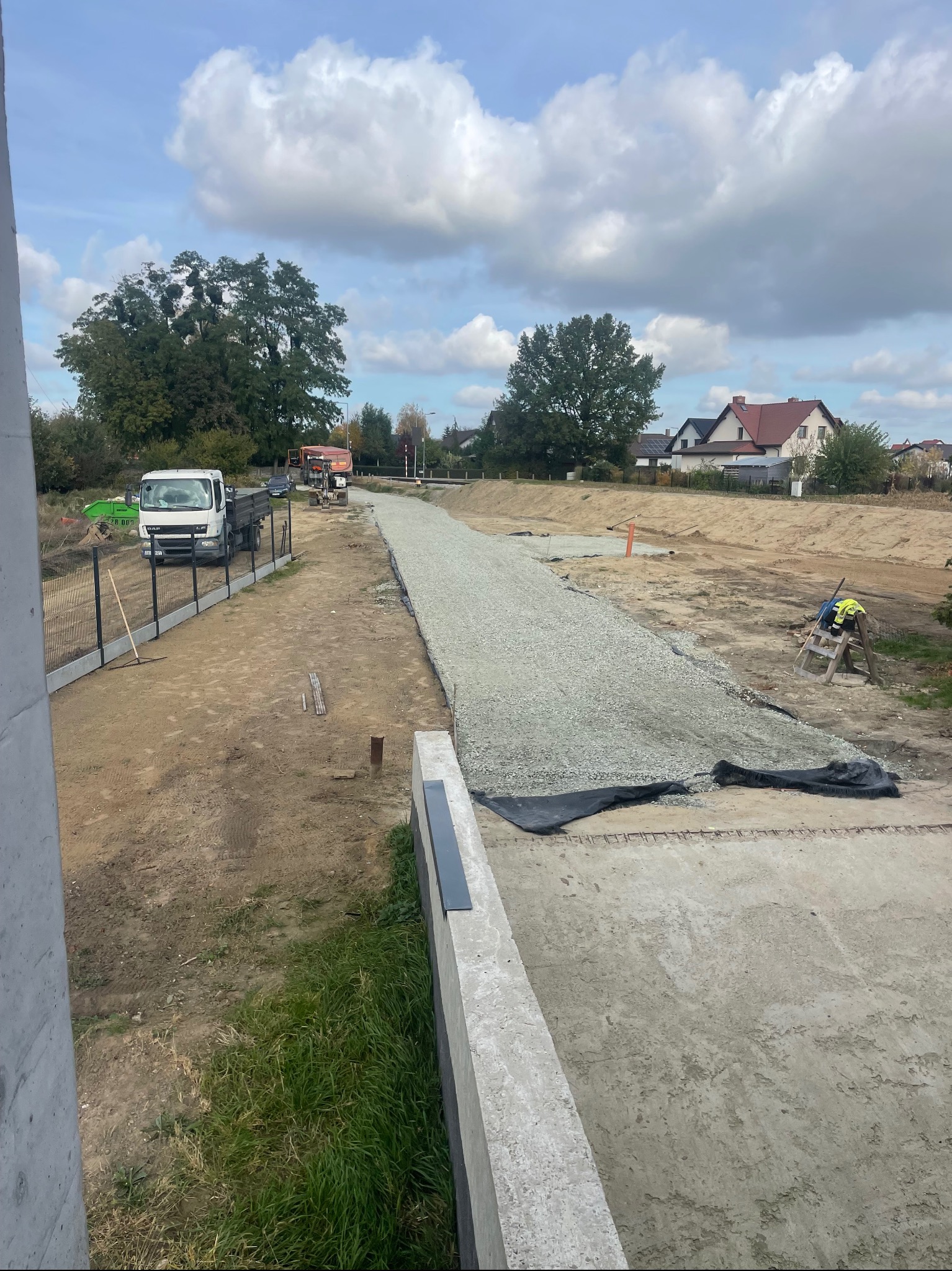 Przygotowanie podłoża pod budowę drogi: warstwa gruzu na czarnej geowłókninie, widoczny ciężarowy samochód i walec w oddali, ogrodzenie z siatki, teren budowy w słoneczny dzień.