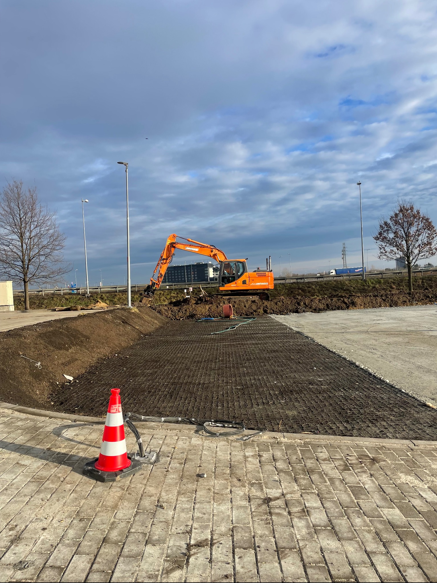 Pomarańczowa koparka Doosan na placu budowy, w tle droga szybkiego ruchu i pochmurne niebo, w pierwszym planie ułożona ażurowa nawierzchnia i pachołek ostrzegawczy.