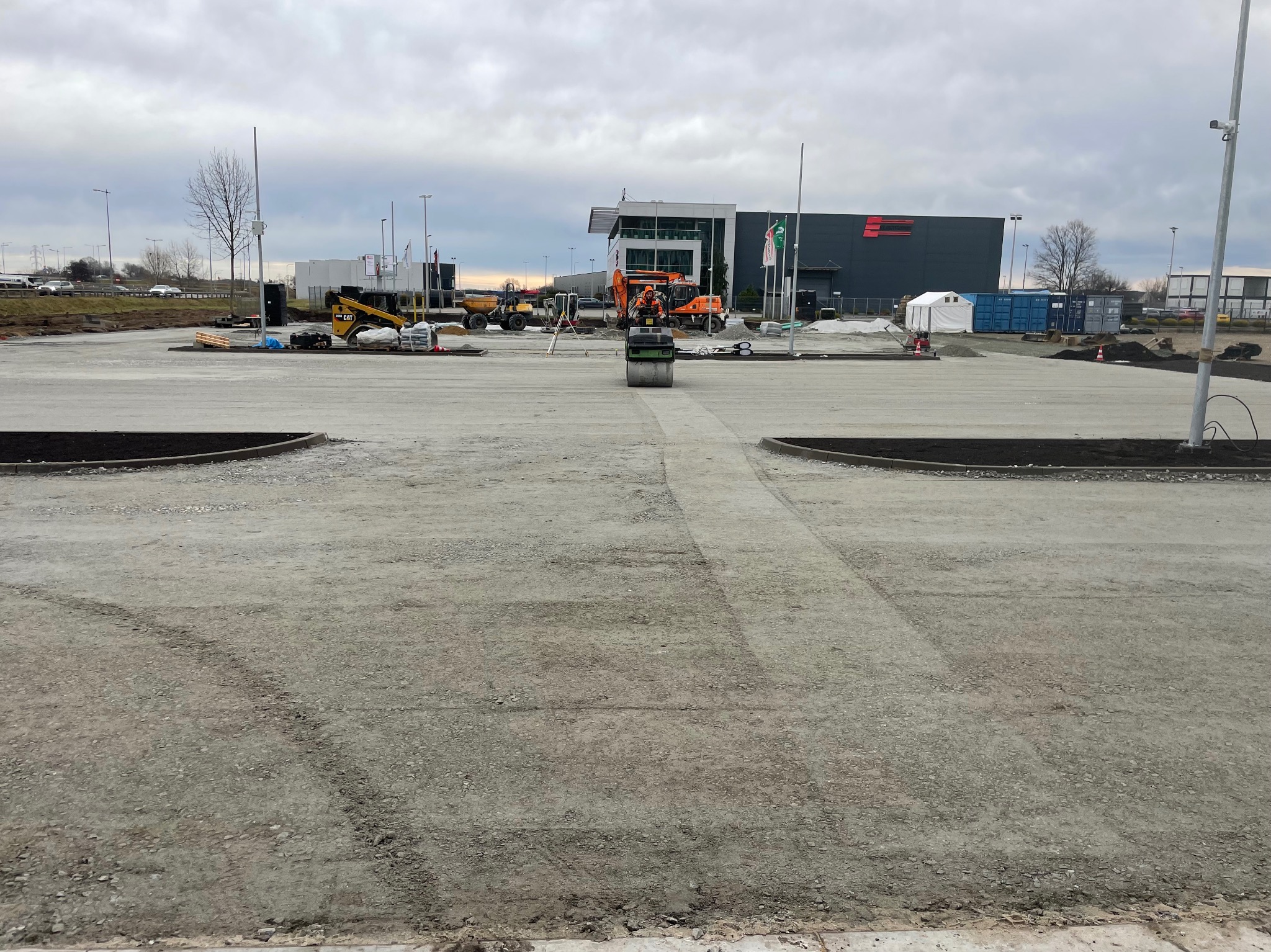 Walec drogowy zagęszcza podłoże na placu budowy, w tle widoczne maszyny budowlane i budynek z logo firmy.