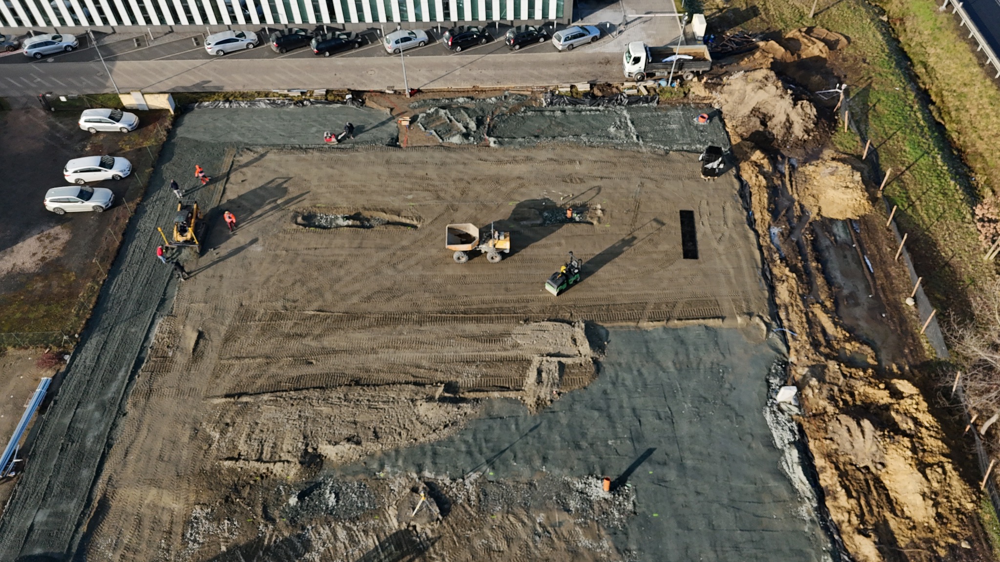 Widok z góry na plac budowy z pracującymi maszynami: walcem, koparką i wywrotką. Część terenu wyrównana i utwardzona, widoczne ślady opon i stopy pracowników w pomarańczowych kamizelkach. W tle...