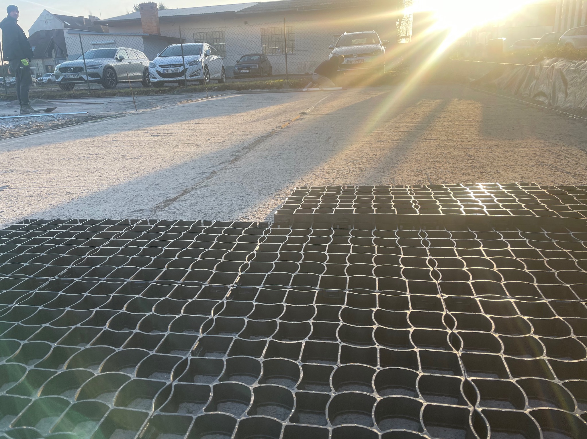 Montaż plastikowych kratek trawnikowych na przygotowanym podłożu, w tle zaparkowane samochody i pracownik wyrównujący teren.